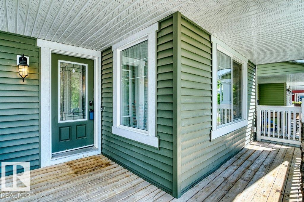 Covered porch with wood flooring, green siding, a decorative glass entry door, white trim, and an exterior wall lantern - 1627 167 Street Sw, Edmonton, AB - Outdoor With Deck Patio Veranda With Exterior