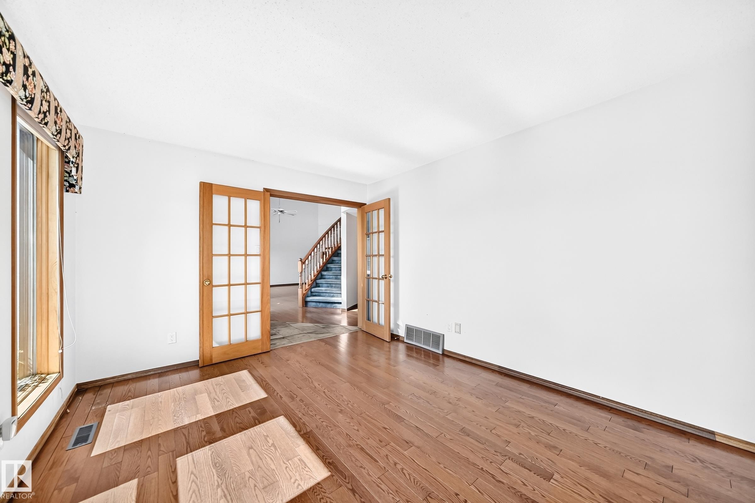Hardwood-finish flooring throughout, featuring a broad window with wood trim, and French doors leading to an interior space with a staircase - 4208 Ramsay Road Nw, Edmonton, AB - Indoor Photo Showing Other Room