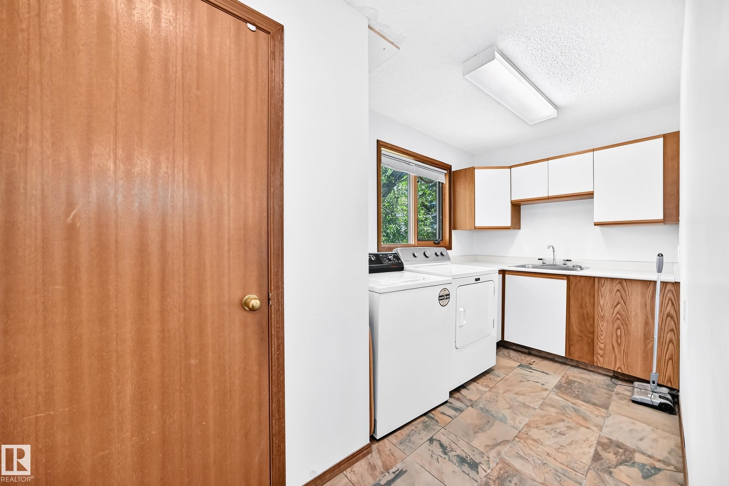 Functional laundry area featuring stone-finish flooring, a window offering natural light, white upper cabinetry, and wood-finish lower cabinetry with a built-in sink - 4208 Ramsay Road Nw, Edmonton, AB - Indoor