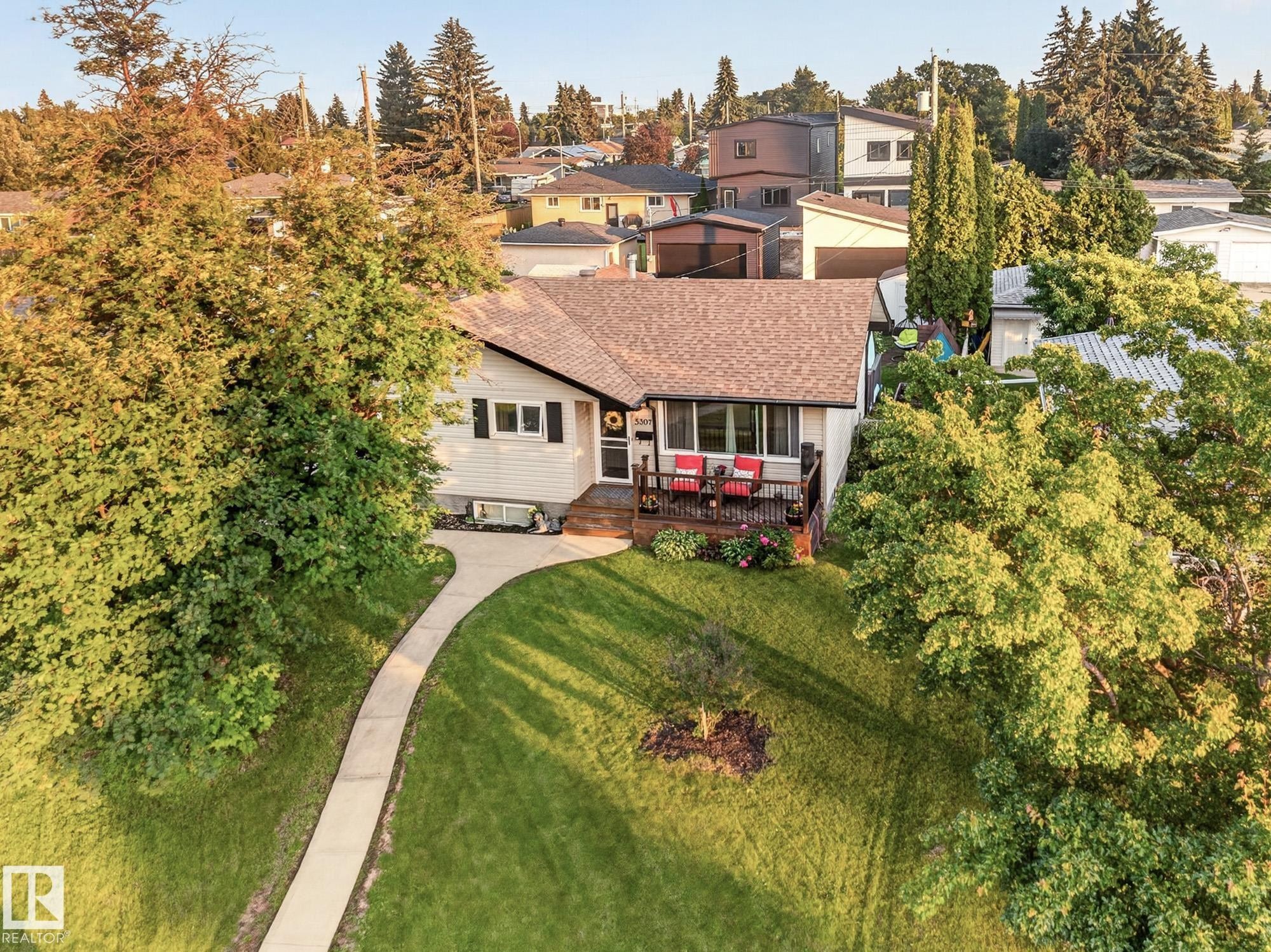 White-sided residence featuring a brown shingle roof, a front deck, a curved concrete walkway, a manicured lawn, and mature trees - 5307 104 Avenue Nw, Edmonton, AB - Outdoor