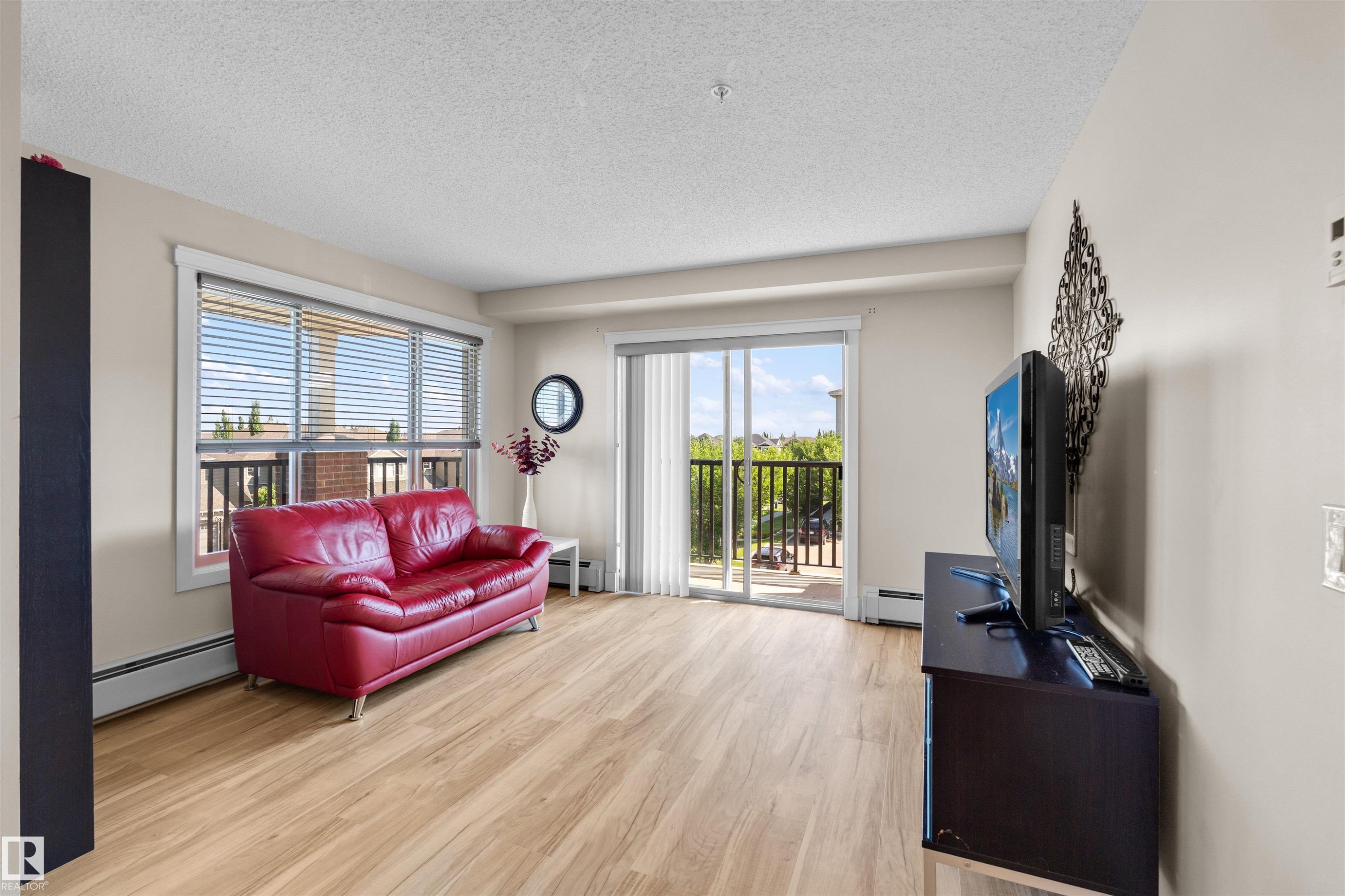 Spacious living area featuring wood-finish flooring, a large window with horizontal blinds, and a sliding glass door leading to an exterior balcony - #317 3719 Whitelaw Ln Nw, Edmonton, AB - Indoor Photo Showing Living Room