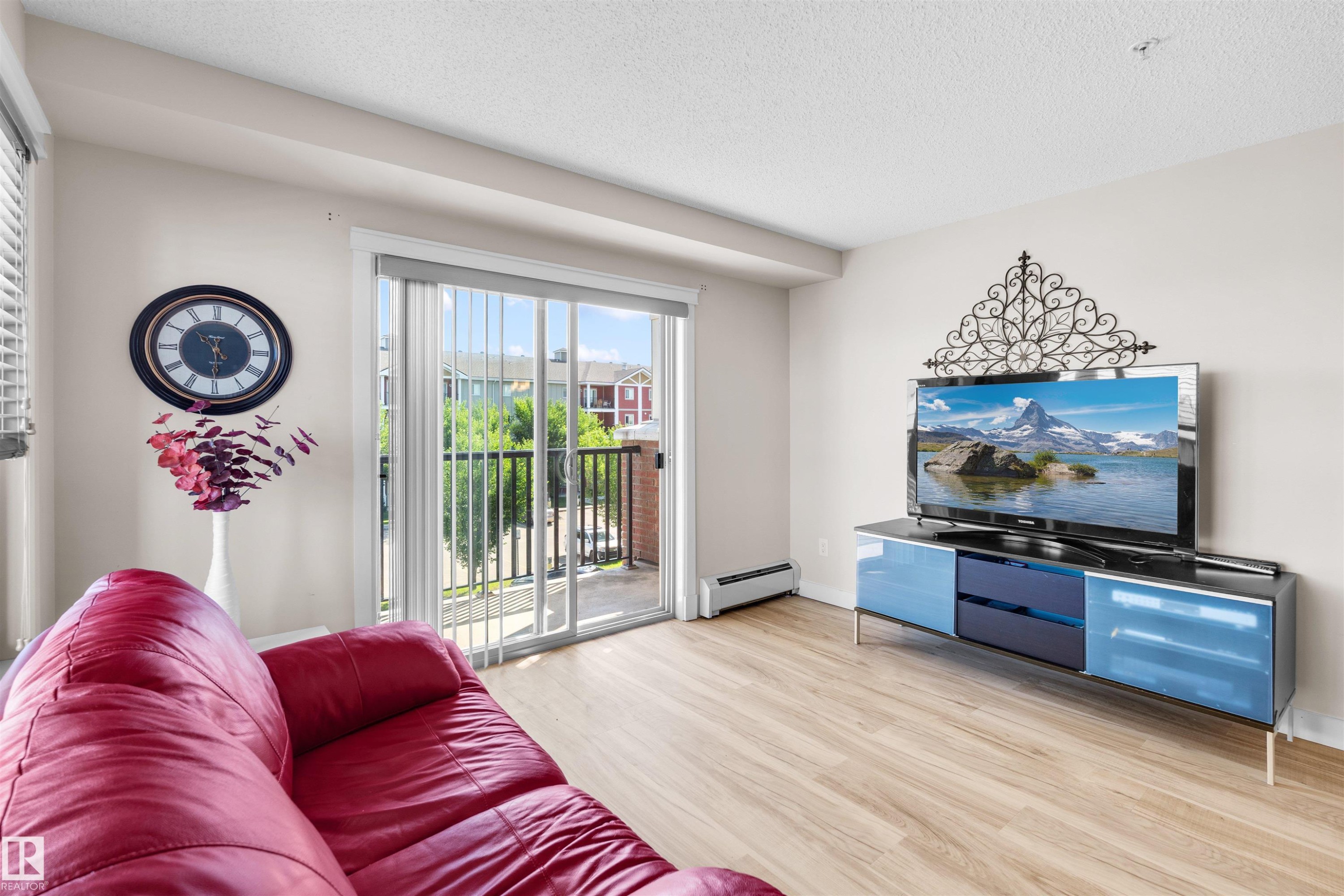 Living area featuring wood-finish flooring, light-toned walls, and sliding glass doors opening to an exterior balcony with black railings - #317 3719 Whitelaw Ln Nw, Edmonton, AB - Indoor Photo Showing Living Room