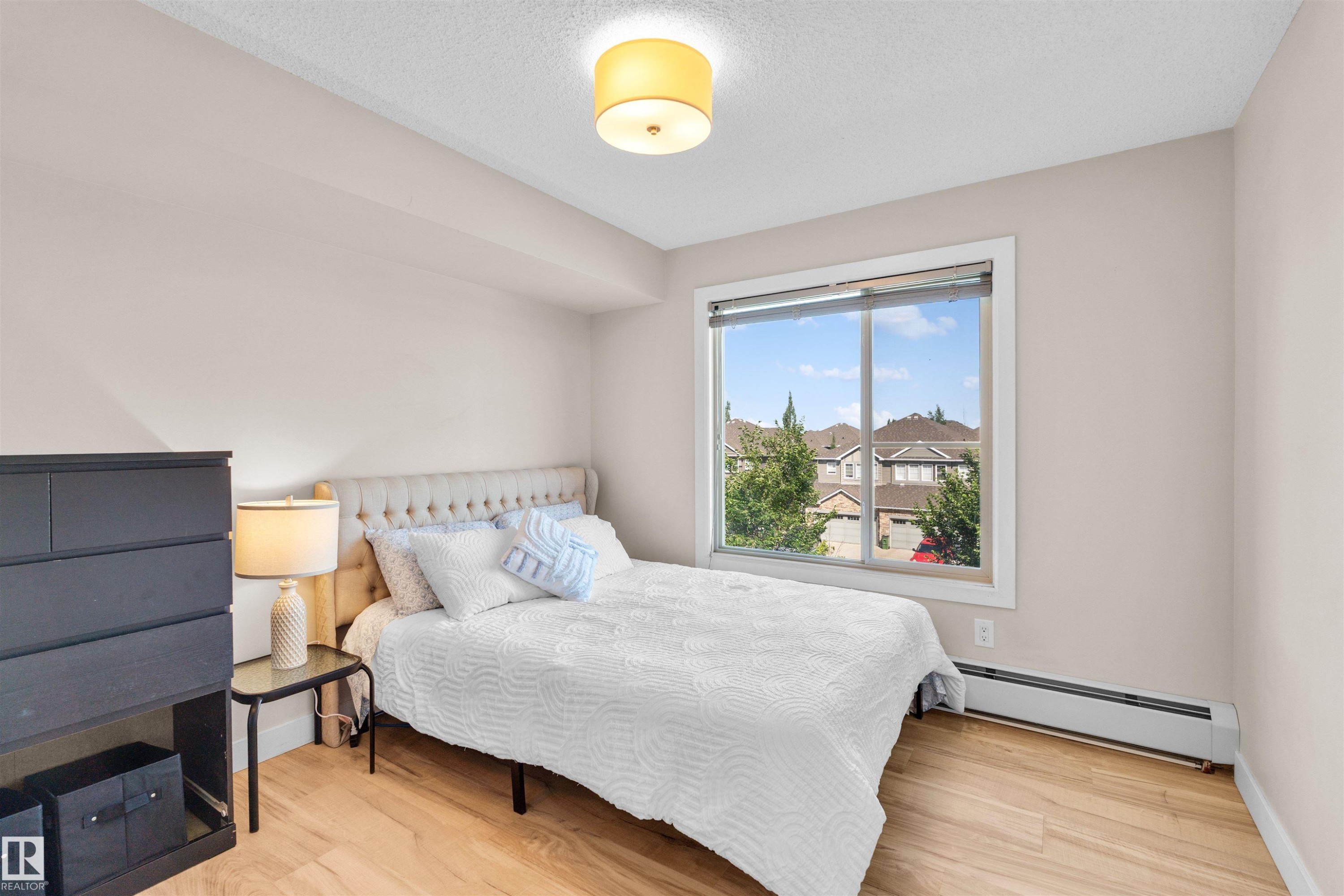 Bedroom featuring light-toned walls, wood-finish flooring, a contemporary ceiling light fixture, a window with white trim, and baseboard heating - #317 3719 Whitelaw Ln Nw, Edmonton, AB - Indoor Photo Showing Bedroom