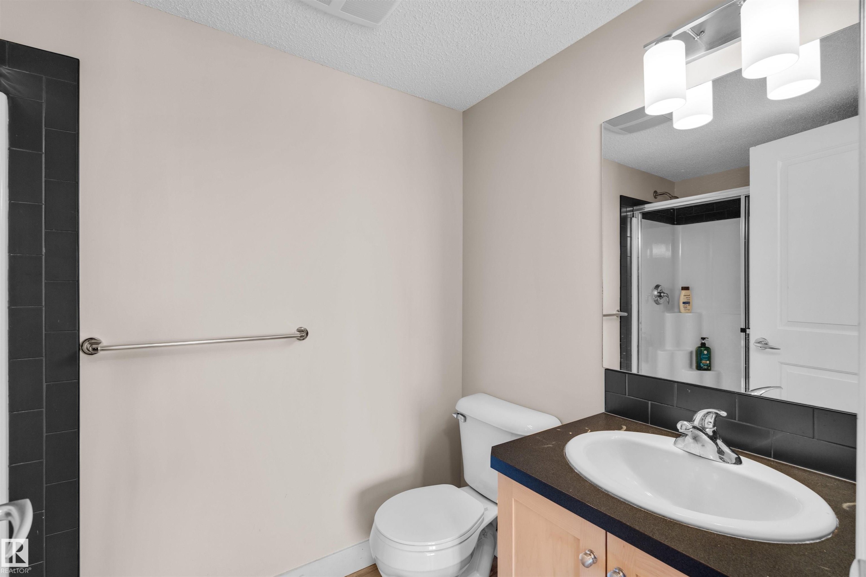 Bathroom featuring a vanity with a drop-in sink, a wall-mounted mirror, overhead lighting, a shower enclosure with a glass door, and dark tile accents - #317 3719 Whitelaw Ln Nw, Edmonton, AB - Indoor Photo Showing Bathroom
