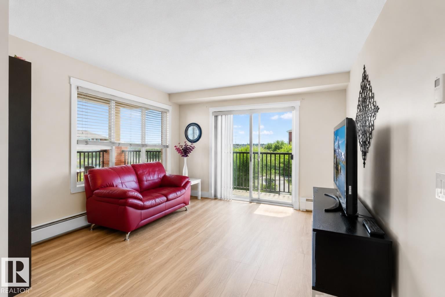 Bright living space featuring wood-finish flooring, a large window with blinds, and a sliding glass door opening to a balcony with black railings - #317 3719 Whitelaw Ln Nw, Edmonton, AB - Indoor Photo Showing Living Room