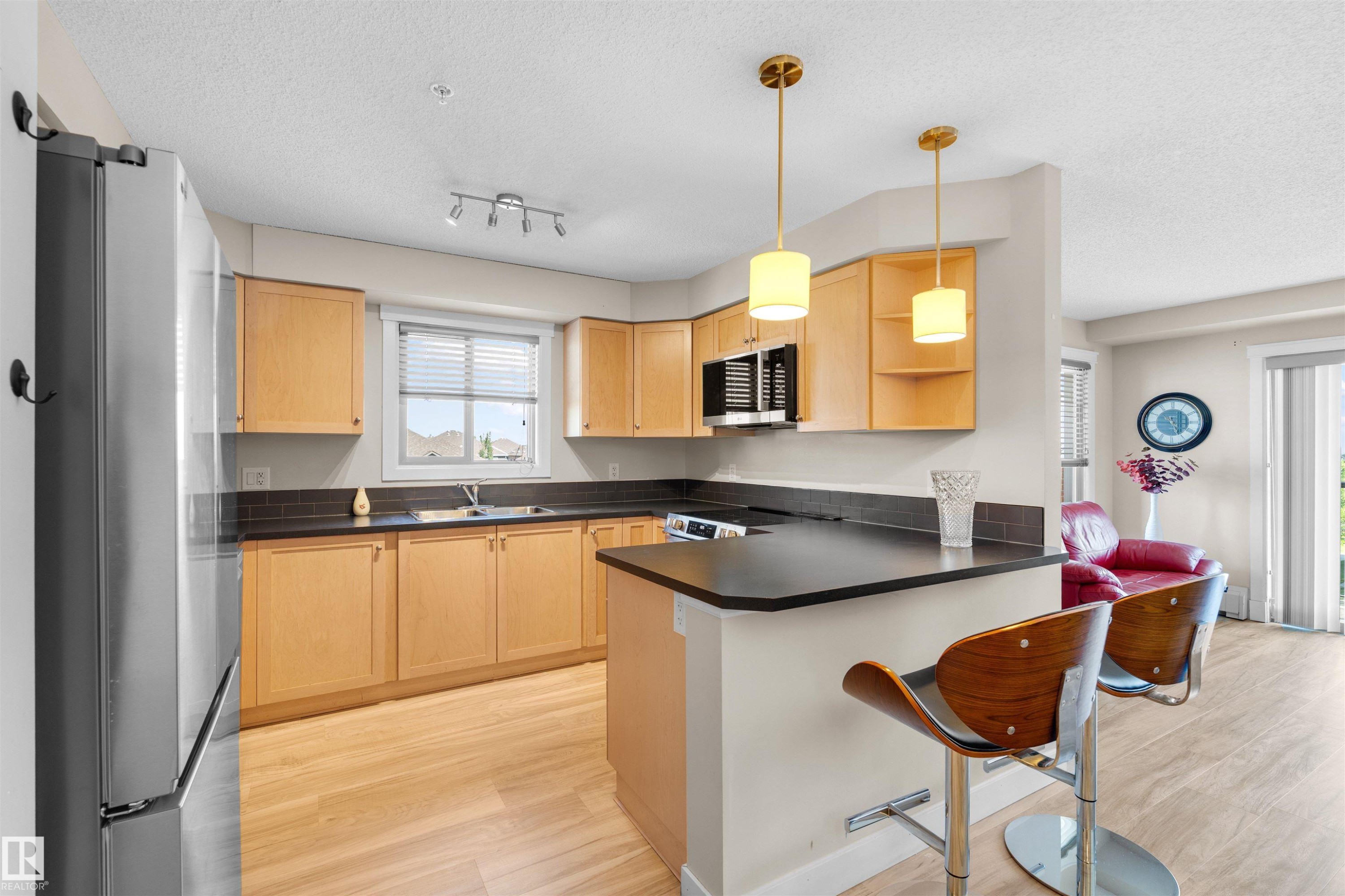 Kitchen featuring light wood-finish cabinetry, dark countertops, and a stainless steel refrigerator - #317 3719 Whitelaw Ln Nw, Edmonton, AB - Indoor Photo Showing Kitchen With Double Sink
