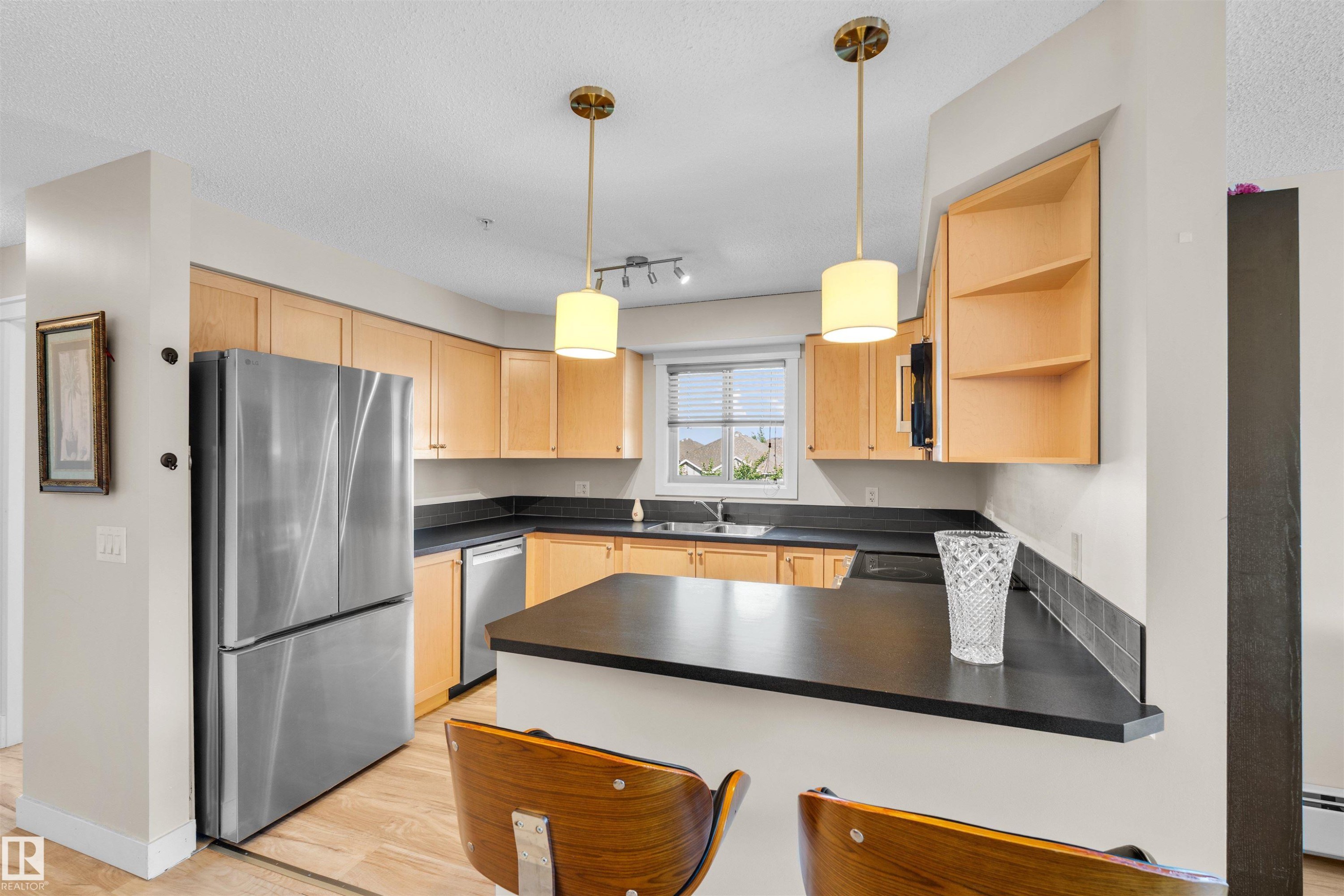Kitchen featuring light wood cabinetry, dark countertops, a stainless steel refrigerator, and recessed lighting - #317 3719 Whitelaw Ln Nw, Edmonton, AB - Indoor Photo Showing Kitchen With Double Sink