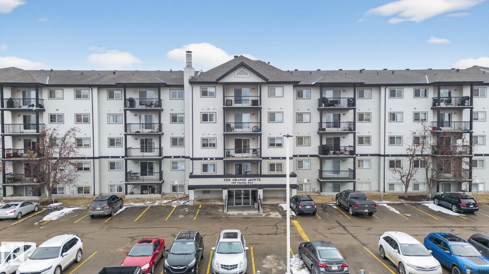 Multi-story residential building featuring balconies with black railings, a central entrance with signage, and a dedicated parking area - 423 2430 Guardian Road, Edmonton, AB - Outdoor With Balcony With Facade