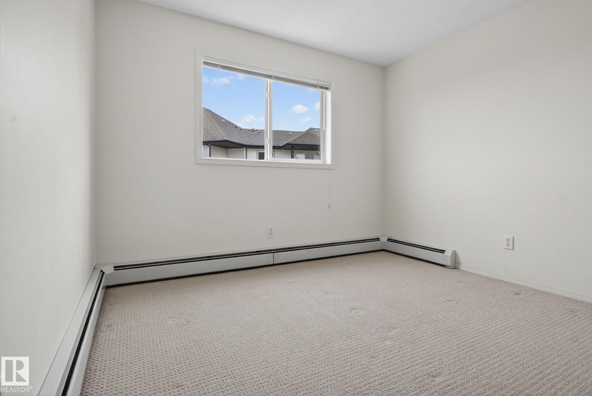 Bright room featuring neutral wall paint, light-colored patterned carpet, a large window, and baseboard heating units - 423 2430 Guardian Road, Edmonton, AB - Indoor Photo Showing Other Room