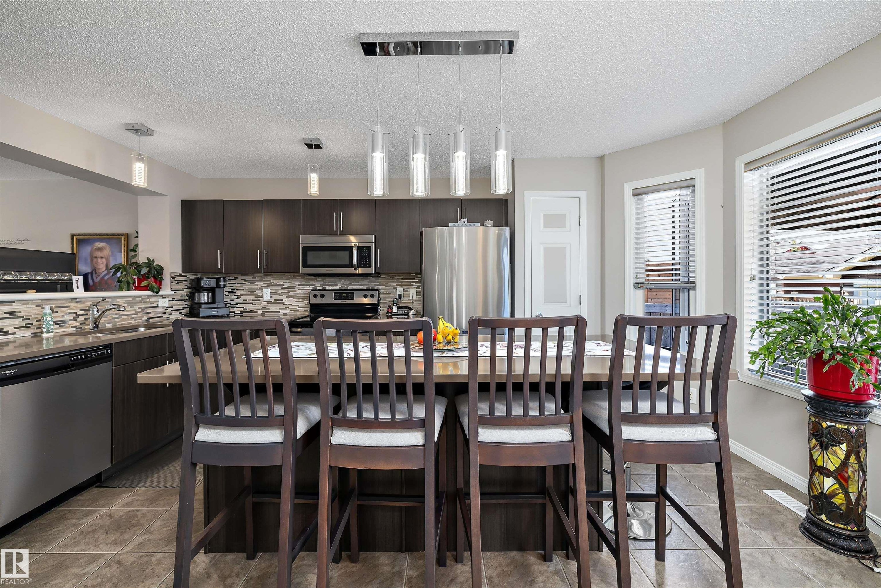 Modern kitchen featuring dark wood-finish cabinetry, stainless steel appliances, a tiled backsplash, and a multi-light pendant fixture over the island - 4688 Crabapple Run Sw, Edmonton, AB - Indoor