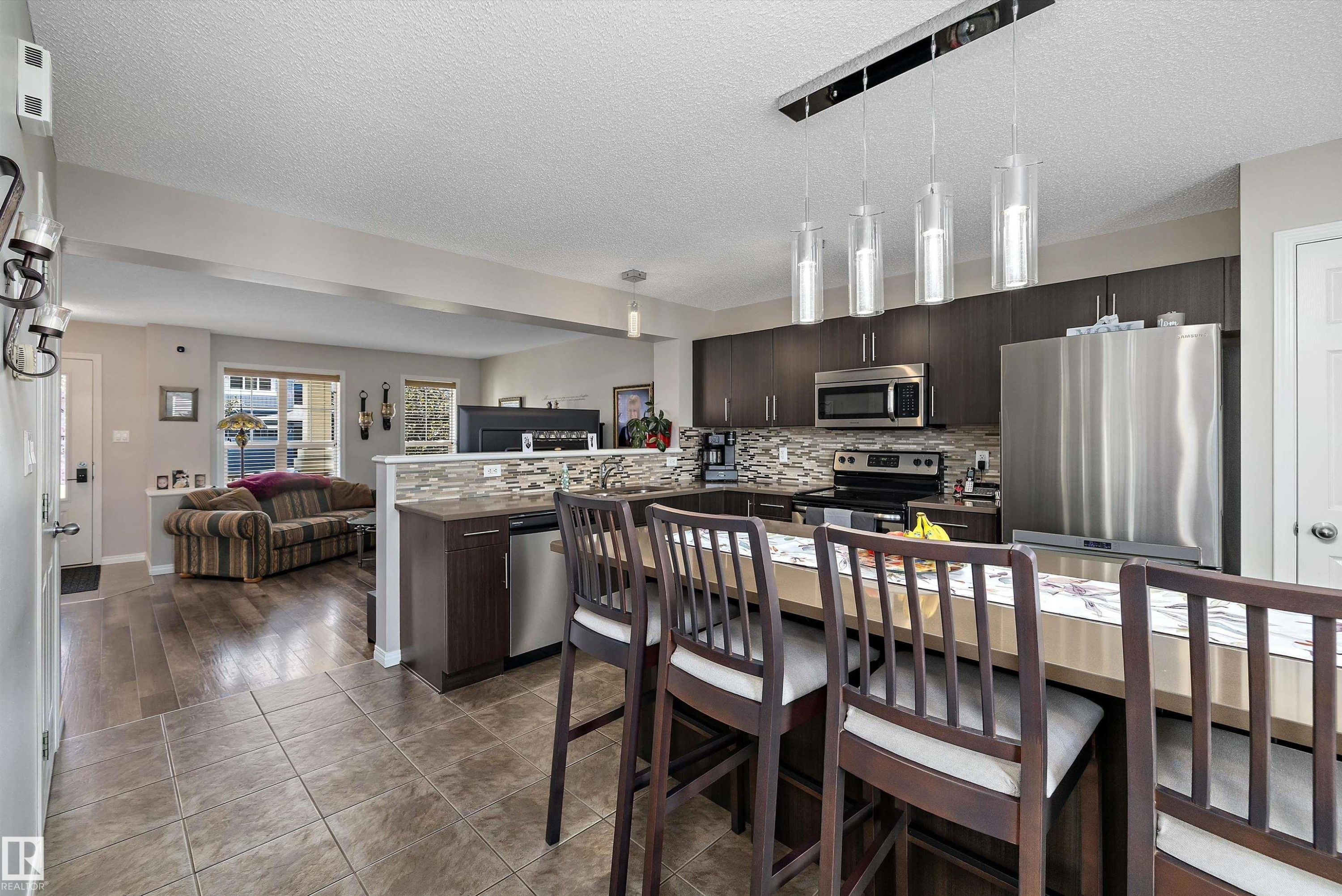 Open-concept kitchen and living area featuring dark wood-finish cabinetry, stainless steel appliances, a tile backsplash, and a breakfast bar island with pendant lighting - 4688 Crabapple Run Sw, Edmonton, AB - Indoor Photo Showing Kitchen With Upgraded Kitchen