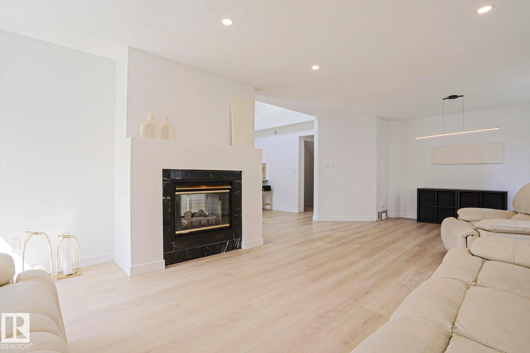 Living area featuring a fireplace with a black marbled surround, light wood-finish flooring, recessed lighting, and a modern linear pendant fixture - 11347 10 Avenue, Edmonton, AB - Indoor Photo Showing Living Room With Fireplace
