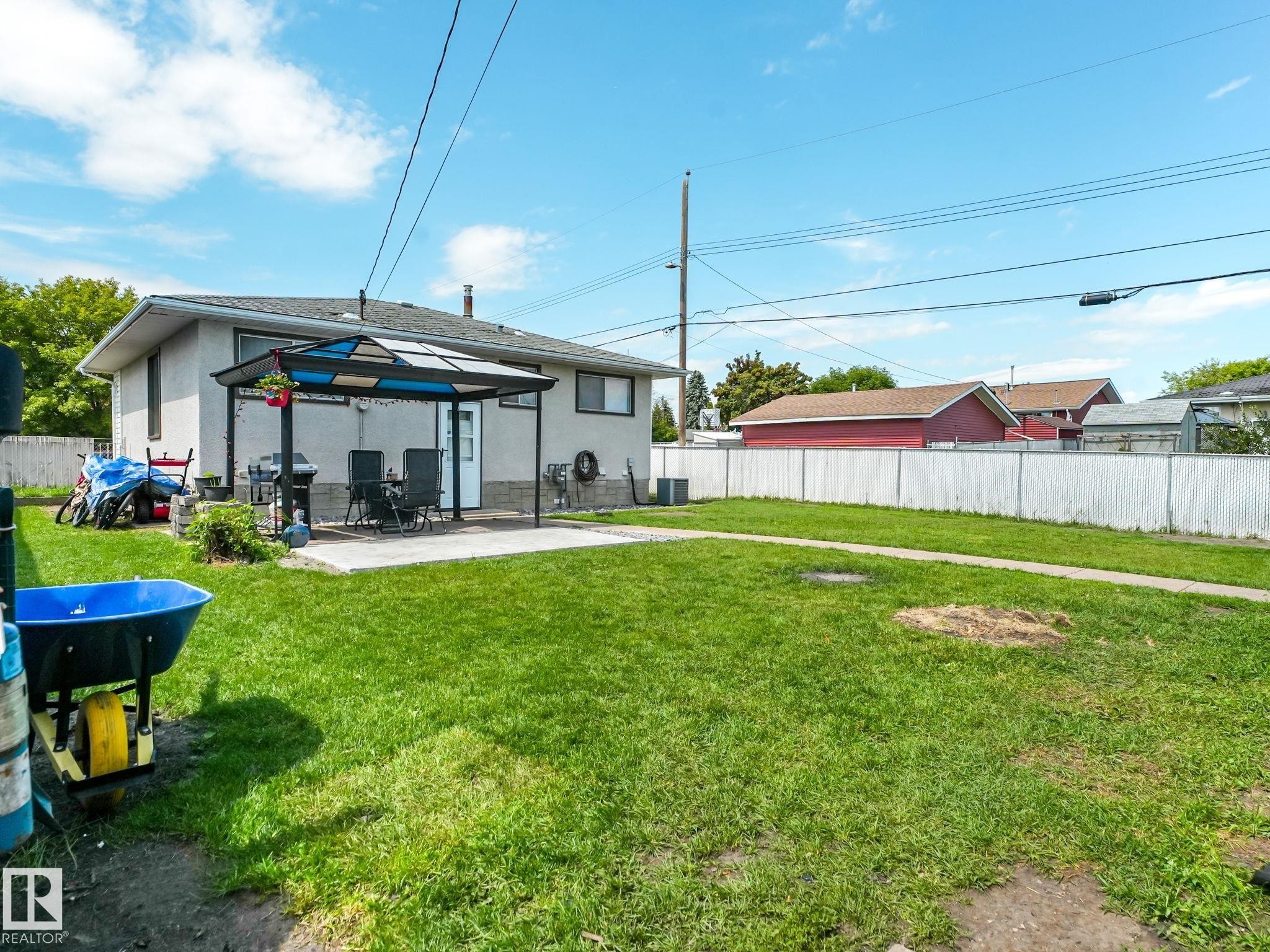 Expansive rear yard featuring a concrete patio with an attached awning, stucco exterior siding, and a white chain-link fence - 13323 58 Street Nw, Edmonton, AB - Outdoor With Backyard