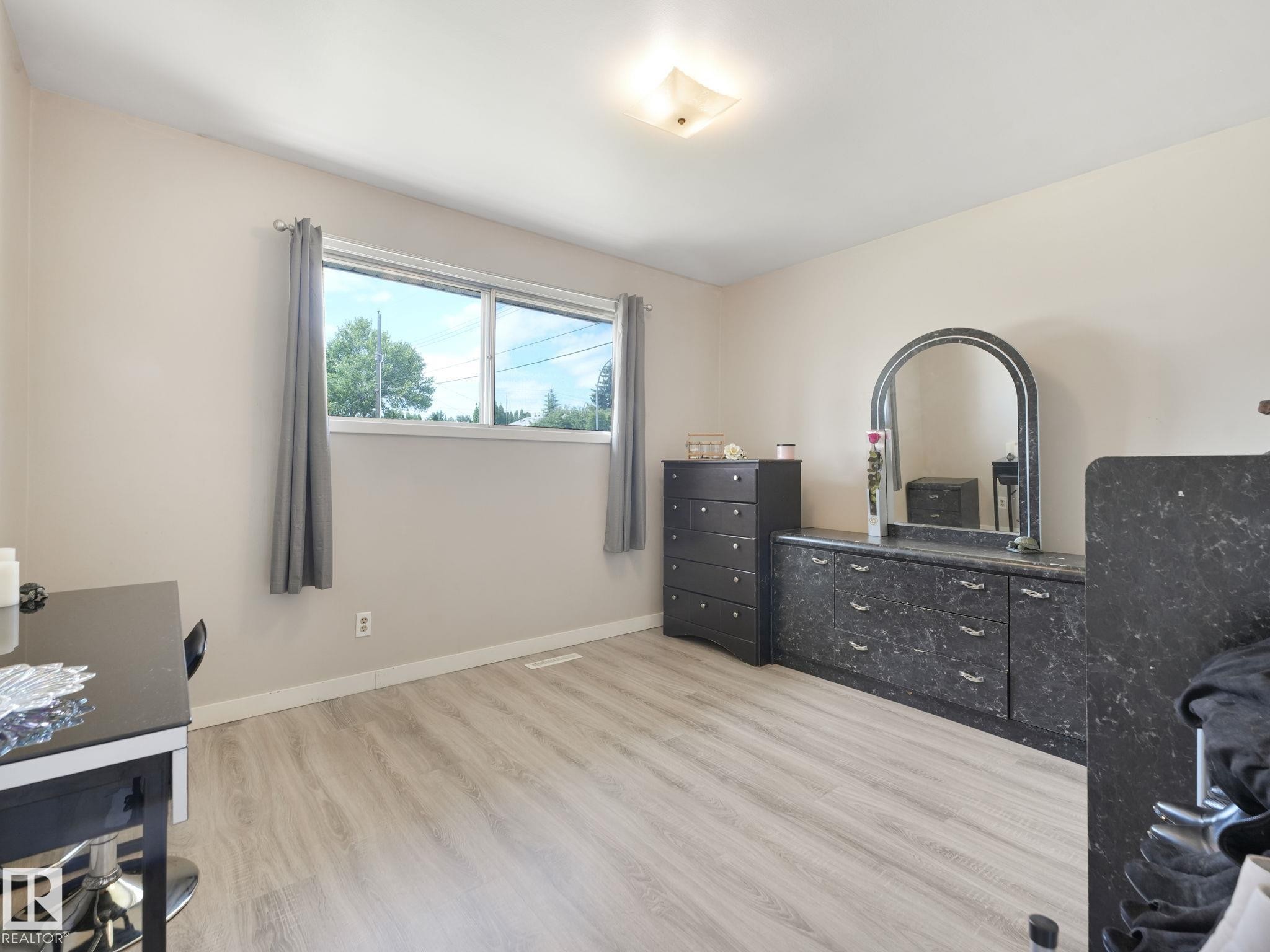 Spacious room featuring light-toned walls, wood-finish flooring, a contemporary ceiling light fixture, and a large window providing natural illumination - 13323 58 Street Nw, Edmonton, AB - Indoor