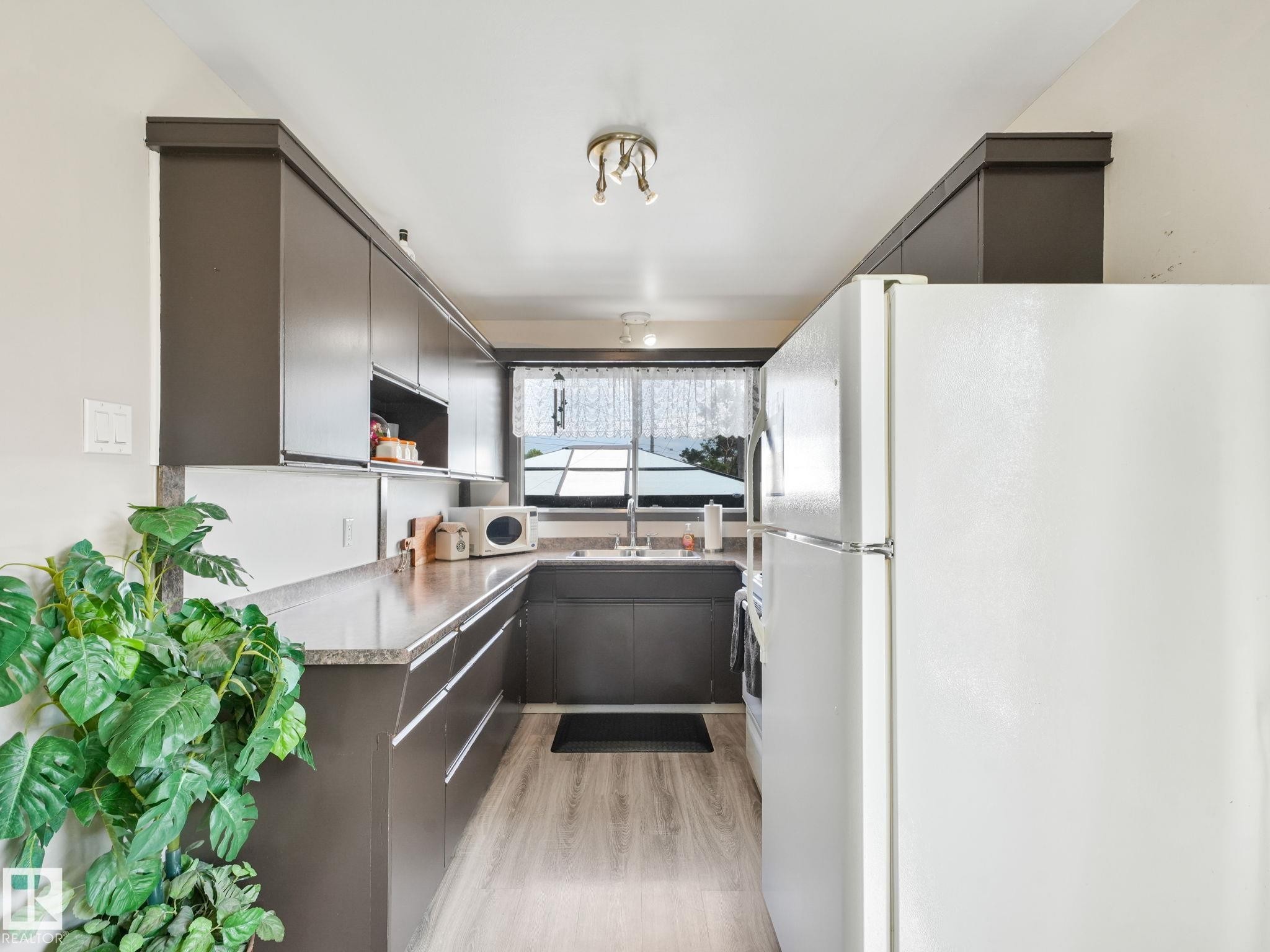 Galley kitchen featuring dark wood-finish cabinetry, light wood-finish flooring, a white refrigerator, a double basin sink, and ample countertop space - 13323 58 Street Nw, Edmonton, AB - Indoor Photo Showing Kitchen