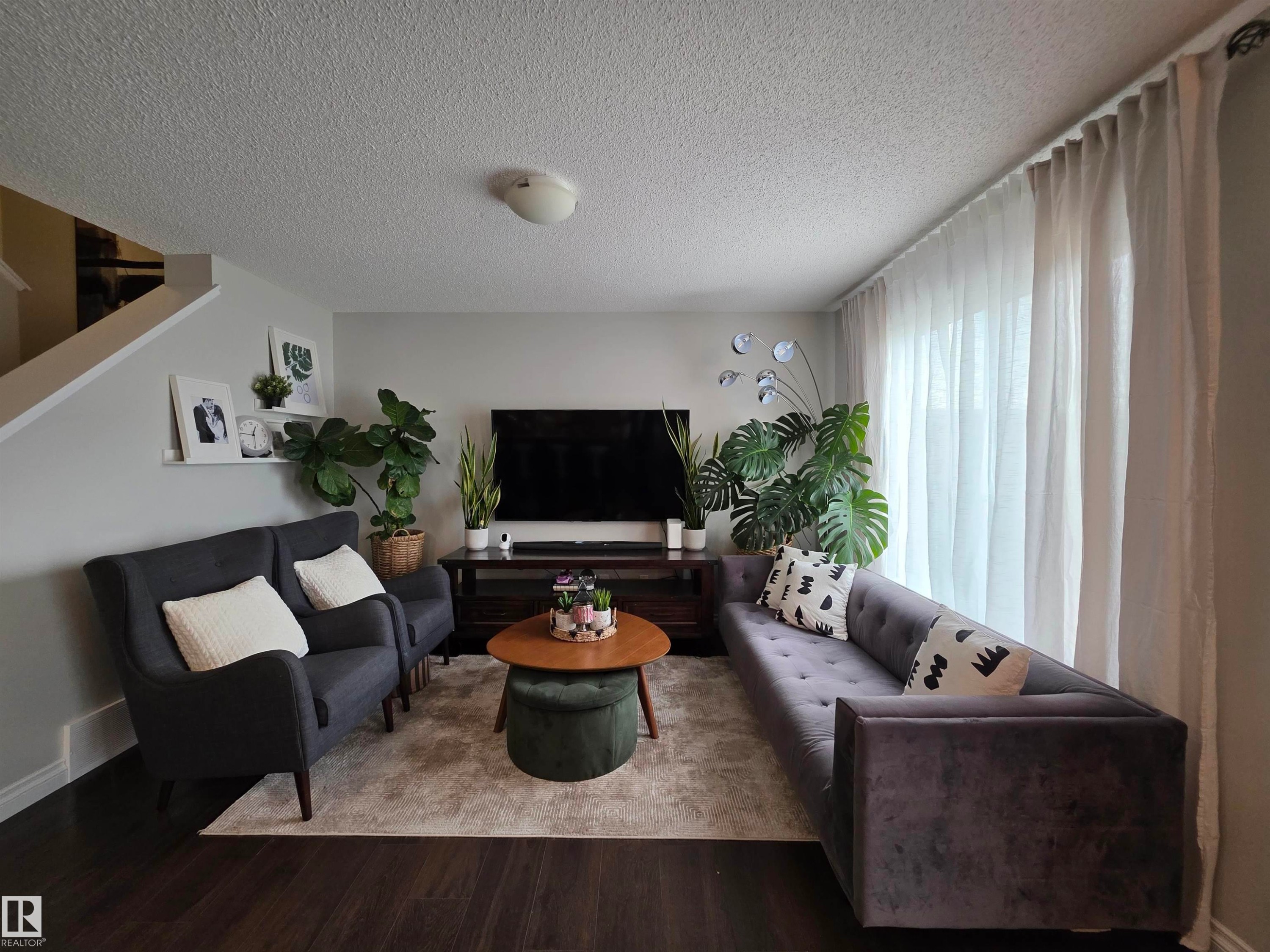 Spacious living area featuring dark wood-finish flooring, a neutral wall color, and a large window with sheer drapes - 491 Ebbers Way, Edmonton, AB