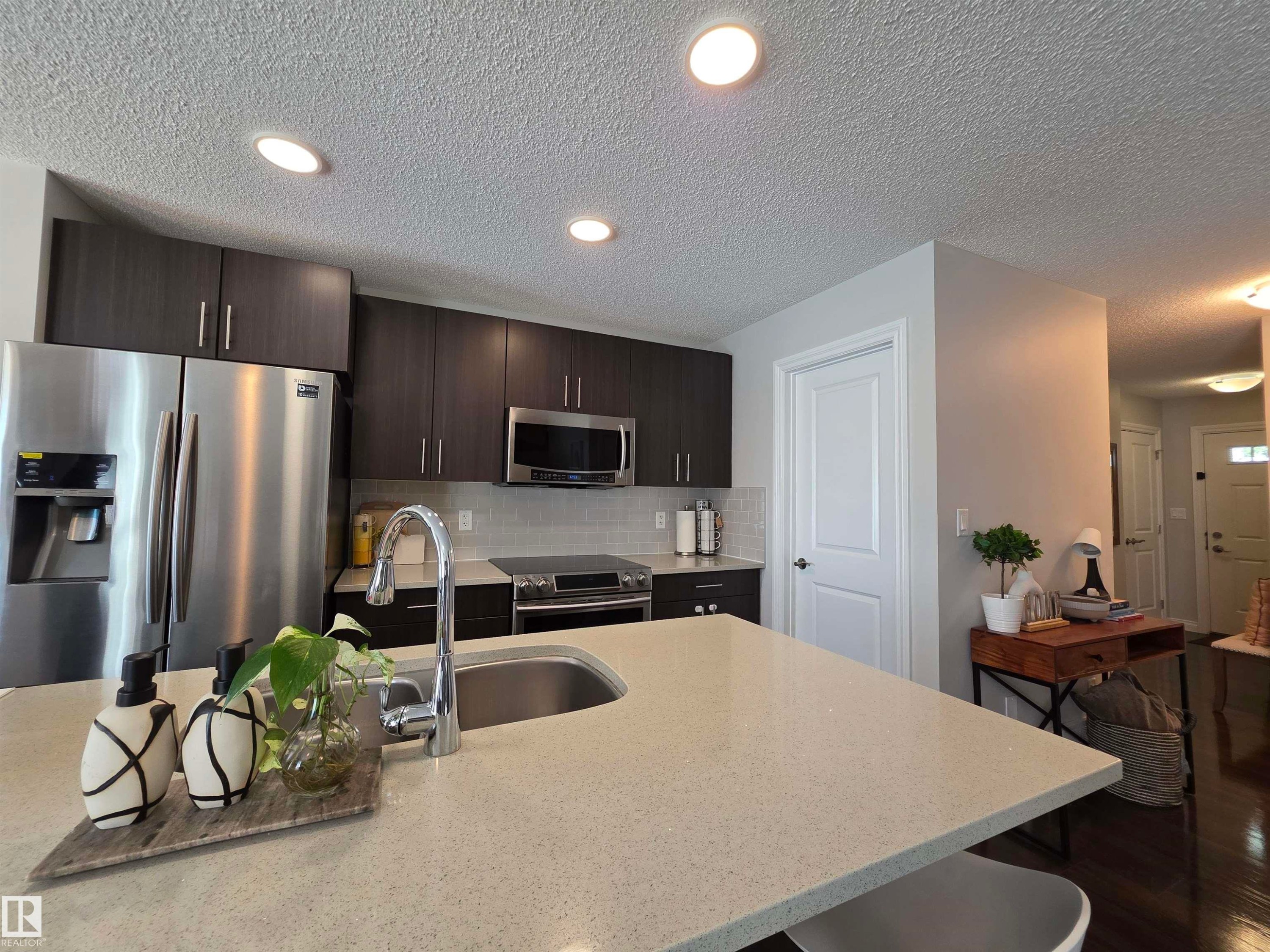 Kitchen featuring a light-toned countertop island with an undermount sink, dark wood-finish cabinetry, stainless steel appliances, and recessed lighting - 491 Ebbers Way, Edmonton, AB