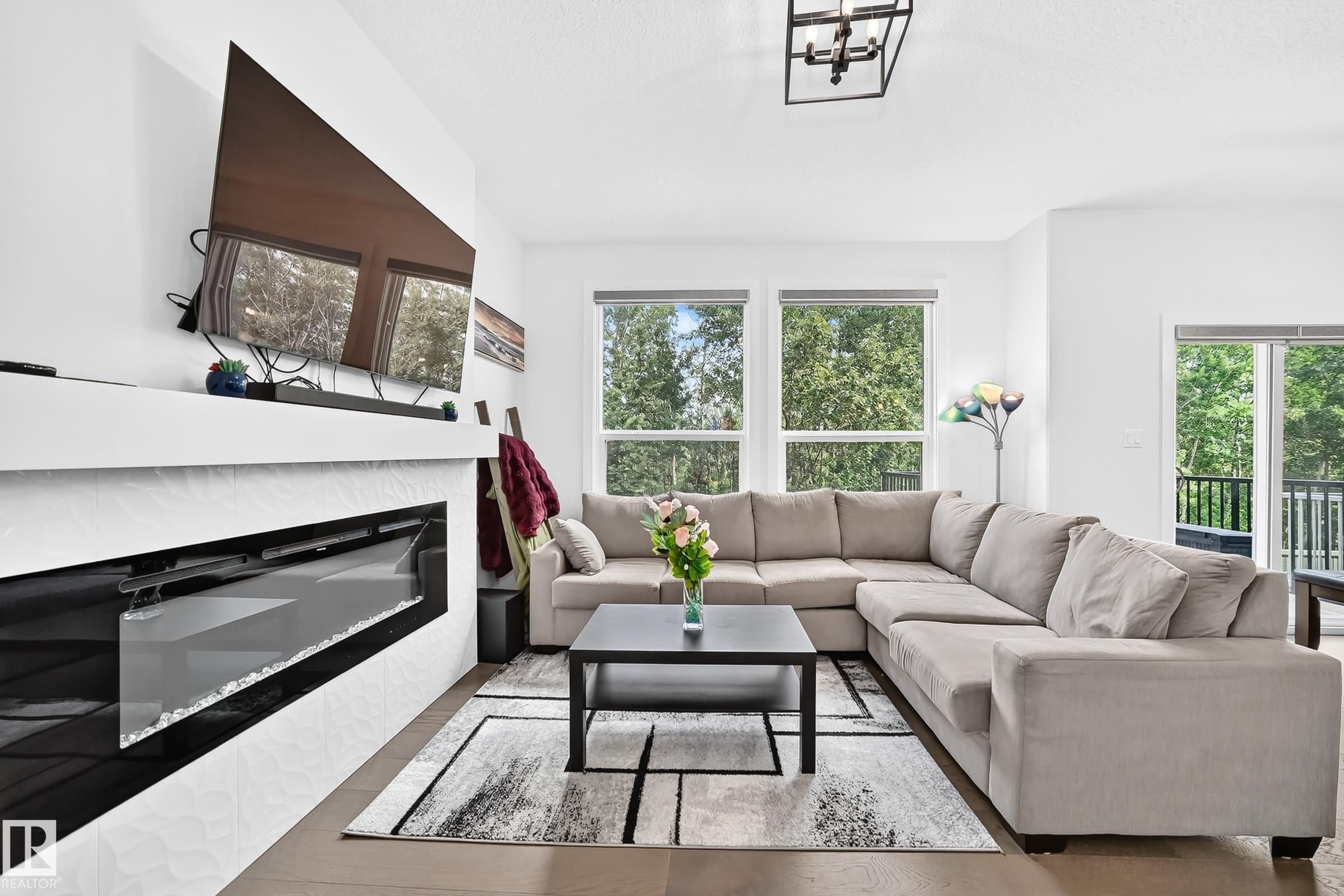 Modern living area featuring a linear electric fireplace with textured white tiling, a wood-finish flooring, and multiple large windows with natural light - 22270 99A Avenue Nw, Edmonton, AB - Indoor Photo Showing Living Room With Fireplace
