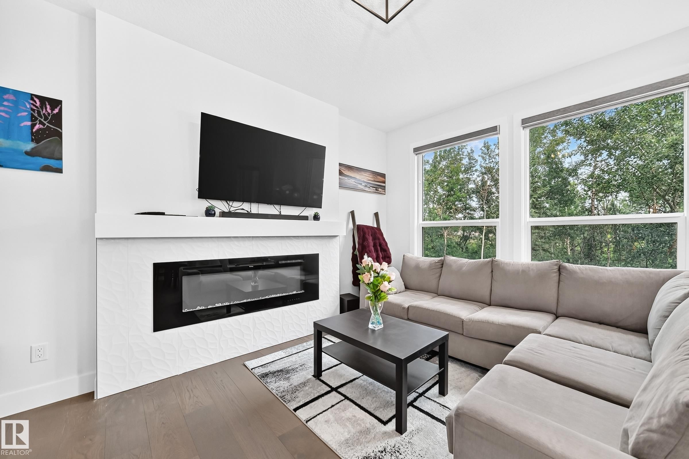 Living area featuring a contemporary linear fireplace with a textured white surround, dark wood-finish flooring, and two large windows offering abundant natural light - 22270 99A Avenue Nw, Edmonton, AB - Indoor Photo Showing Living Room With Fireplace