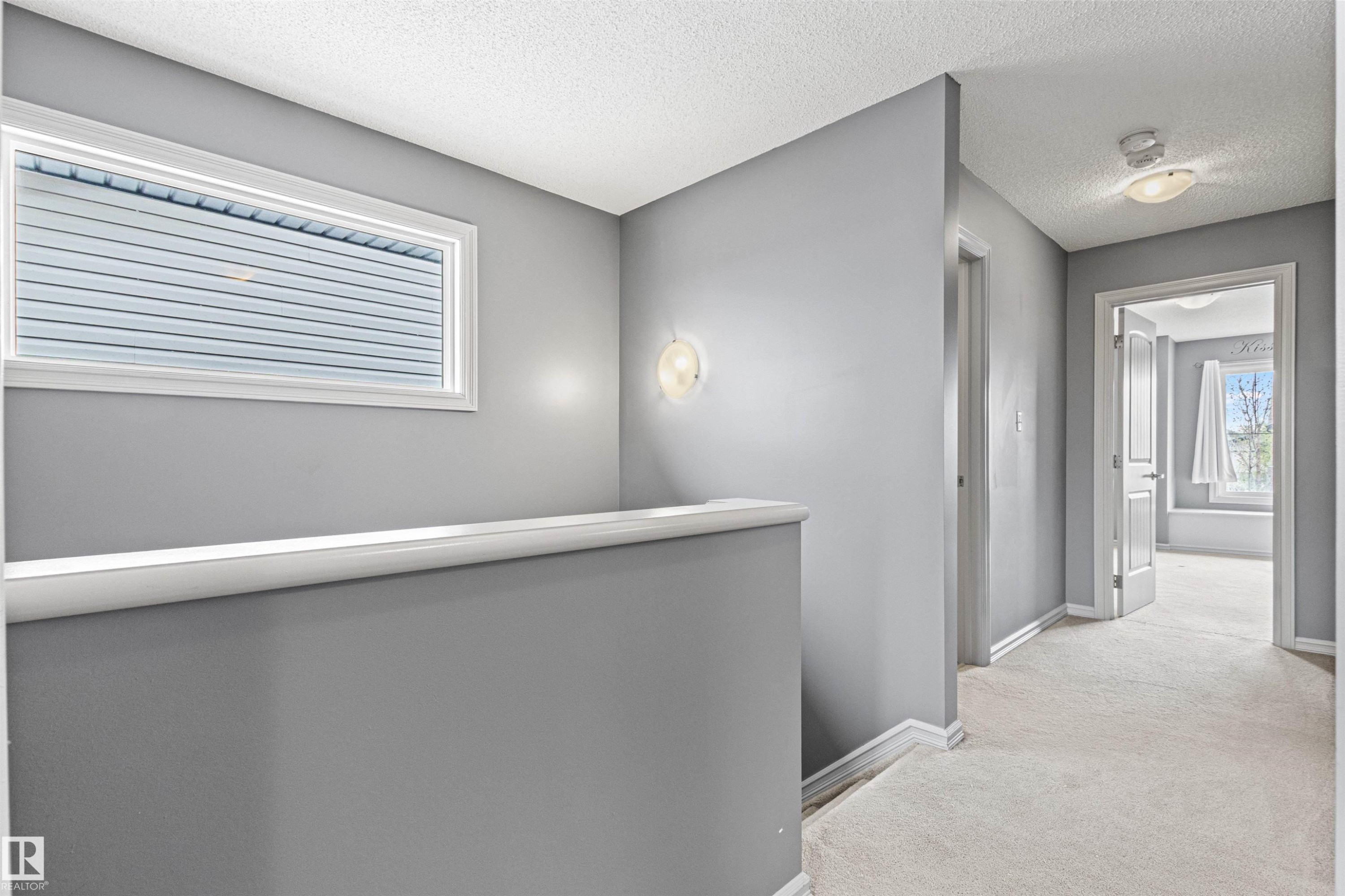 Carpeted hallway featuring a rectangular window with horizontal blinds, a wall sconce, and white trim - 1154 35A Avenue Nw, Edmonton, AB - Indoor Photo Showing Other Room
