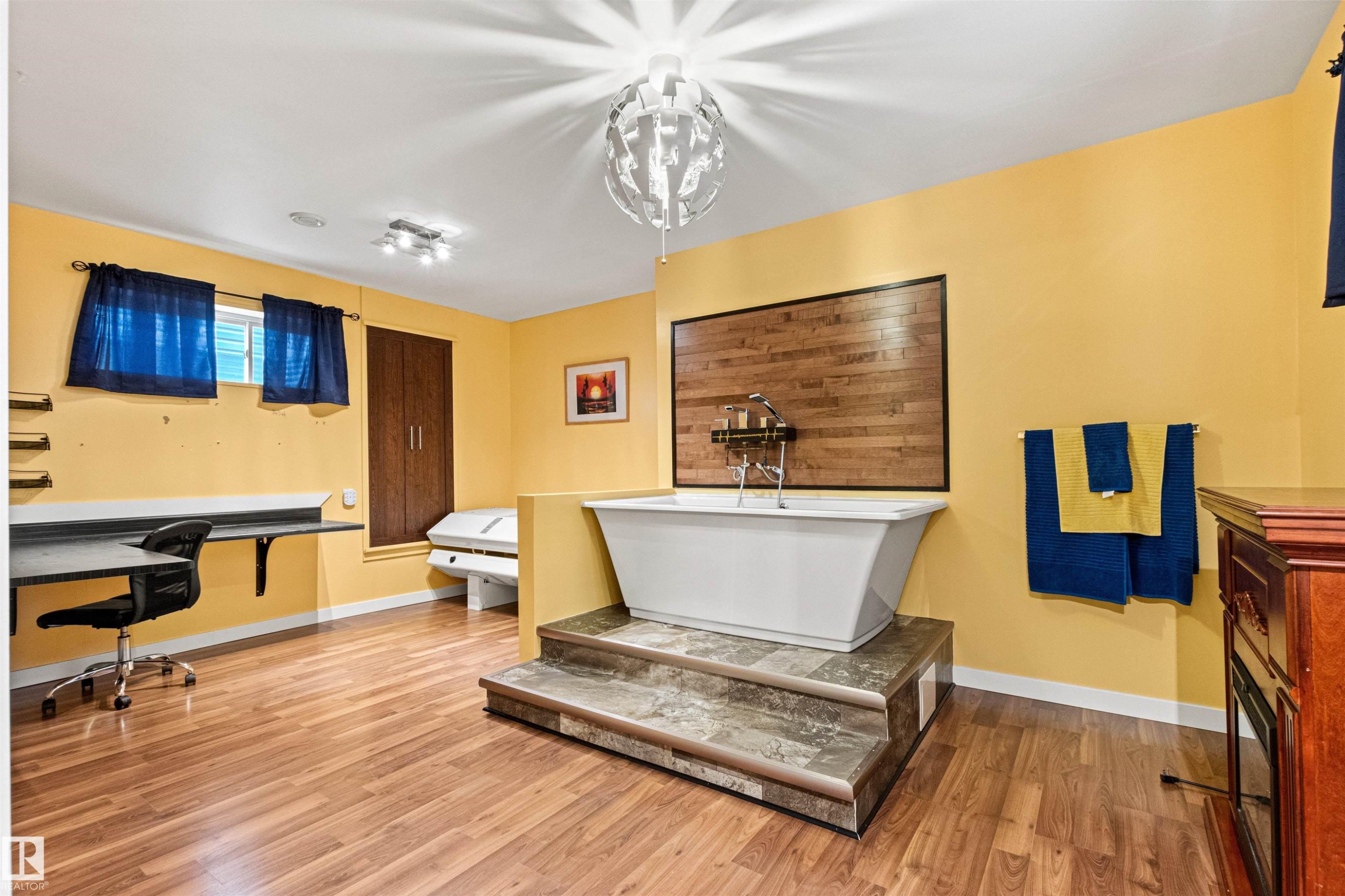 Freestanding bathtub elevated on a two-step platform, featuring a wall-mounted wood-finish accent panel with a chrome faucet and showerhead - 1154 35A Avenue Nw, Edmonton, AB - Indoor