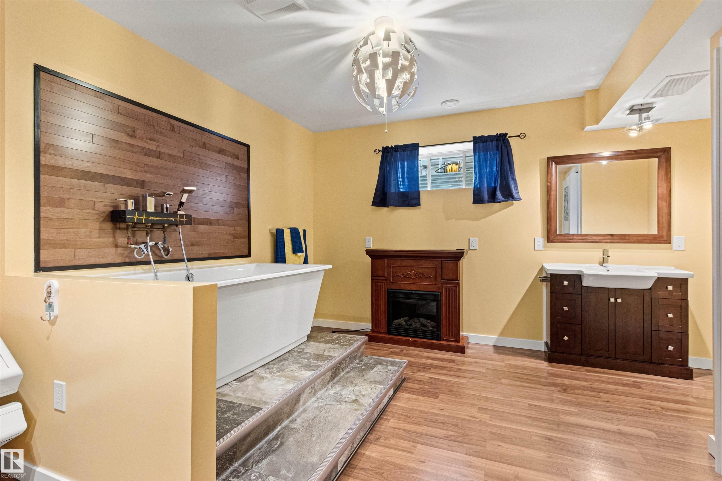 Bathroom featuring a freestanding white tub with a wood-panel accent wall, warm yellow paint, wood-finish flooring, a vanity with a rectangular sink, and a decorative fireplace - 1154 35A Avenue Nw, Edmonton, AB - Indoor With Fireplace