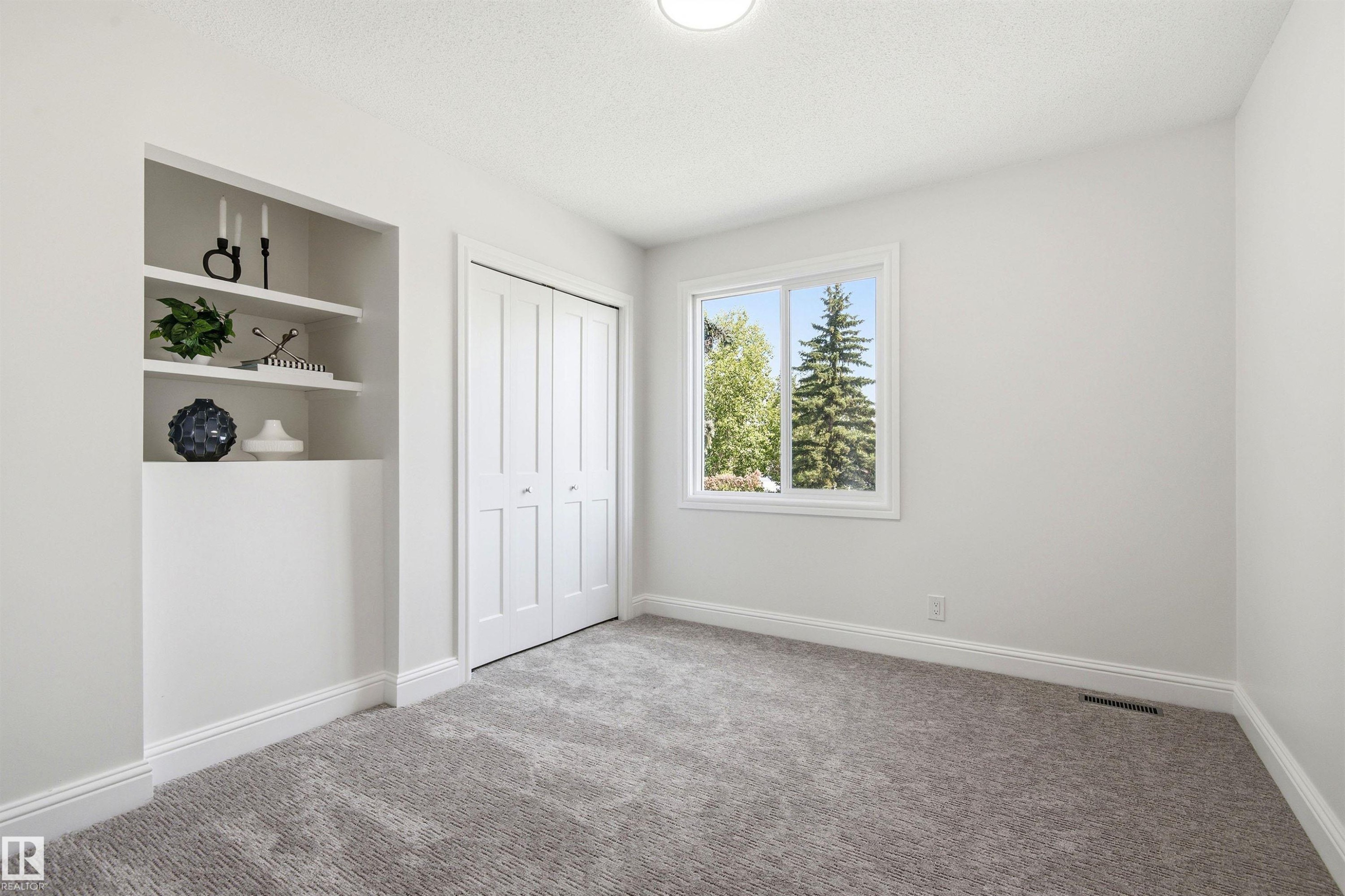 Neutral-toned room featuring a window with exterior tree views, built-in shelving, white bi-fold closet doors, and plush gray carpet - 14335 47 Avenue Nw, Edmonton, AB - Indoor Photo Showing Other Room