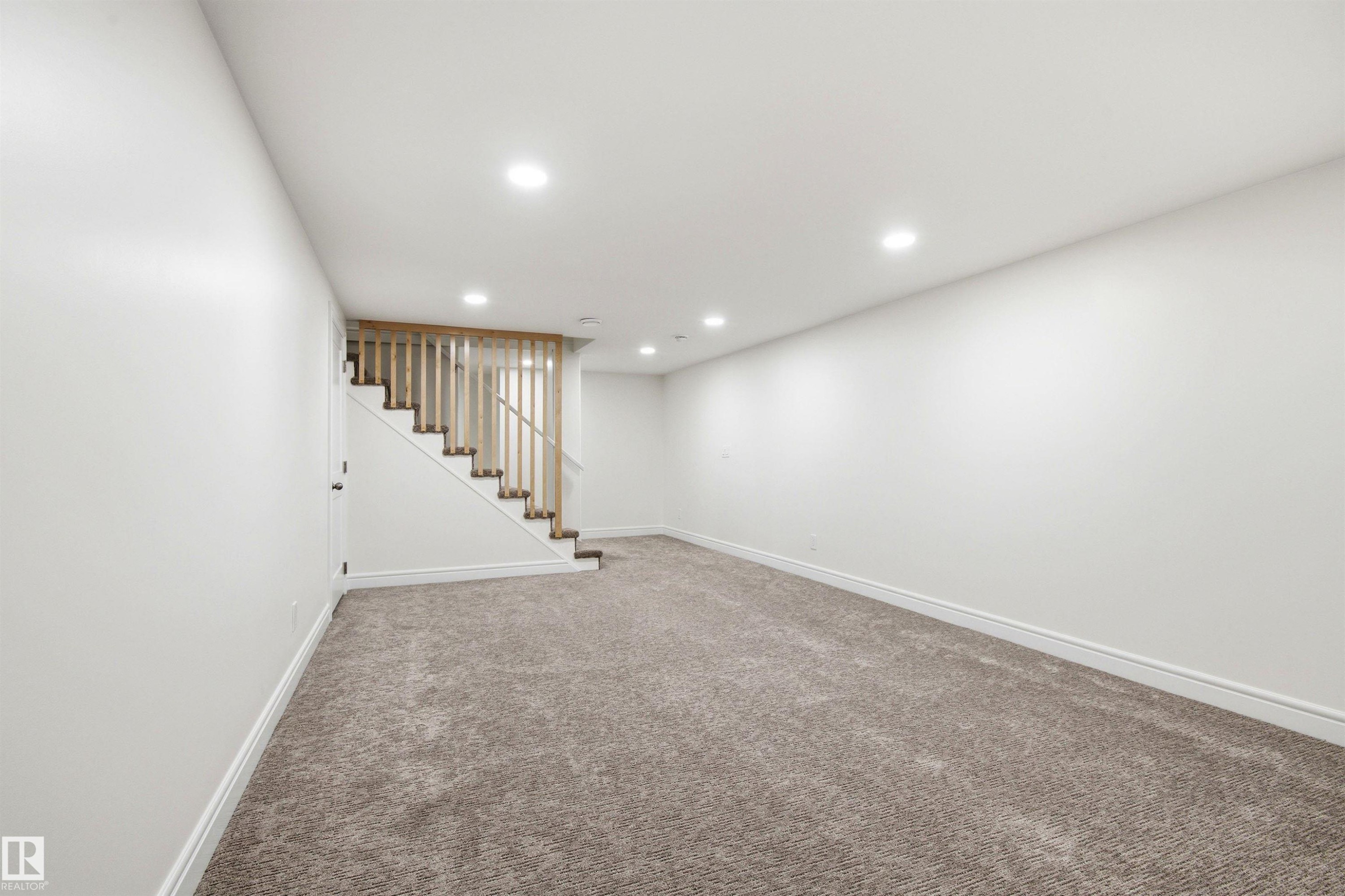 Spacious carpeted area featuring a natural wood-finish staircase with coordinating balusters, recessed ceiling lighting, crisp white walls, and matching baseboards - 14335 47 Avenue Nw, Edmonton, AB - Indoor Photo Showing Other Room