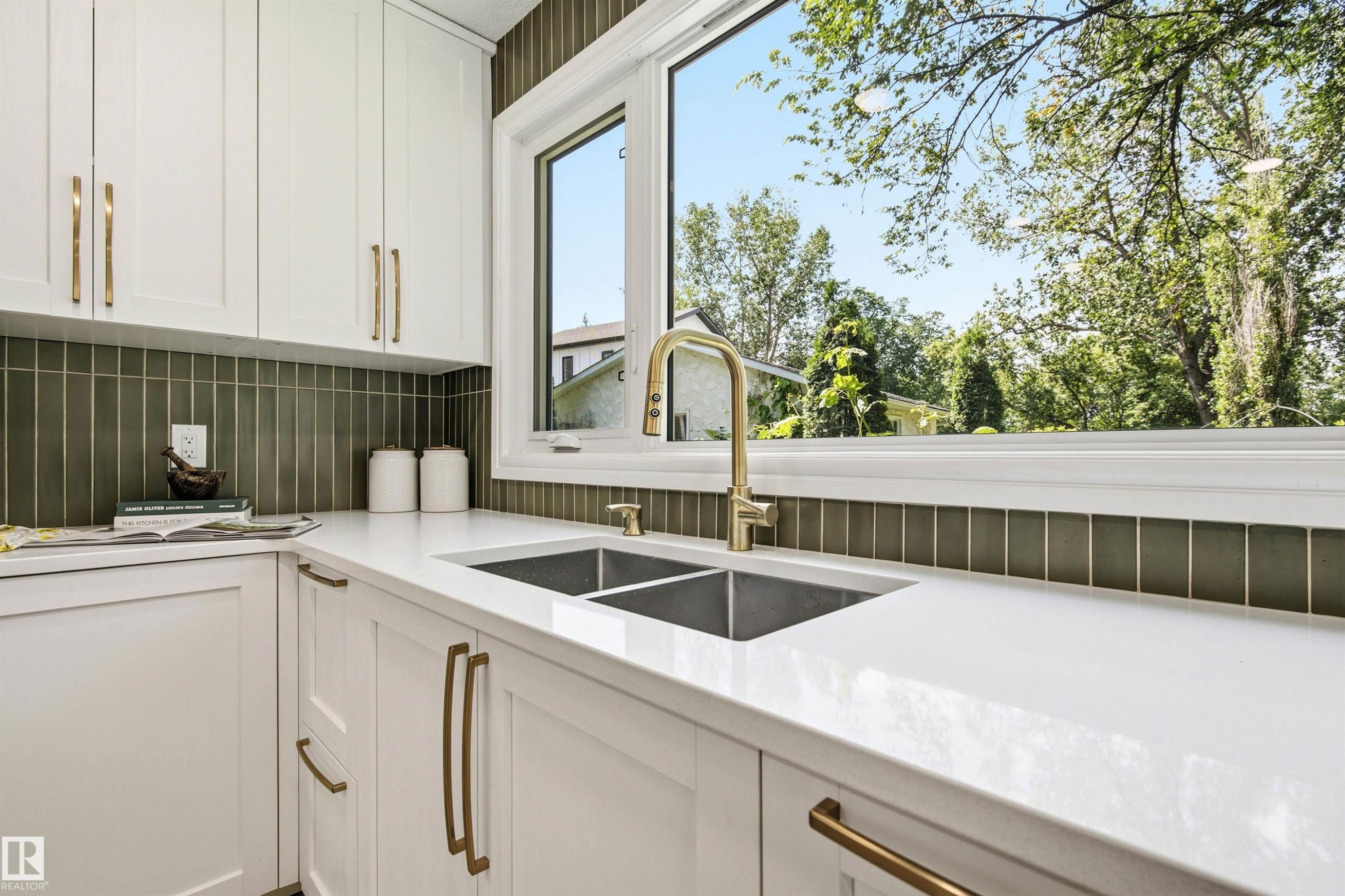 Kitchen featuring white shaker cabinetry, white countertops, a dual basin undermount sink with a brass-finish faucet, and an olive green vertical tile backsplash - 14335 47 Avenue Nw, Edmonton, AB - Indoor Photo Showing Kitchen With Double Sink