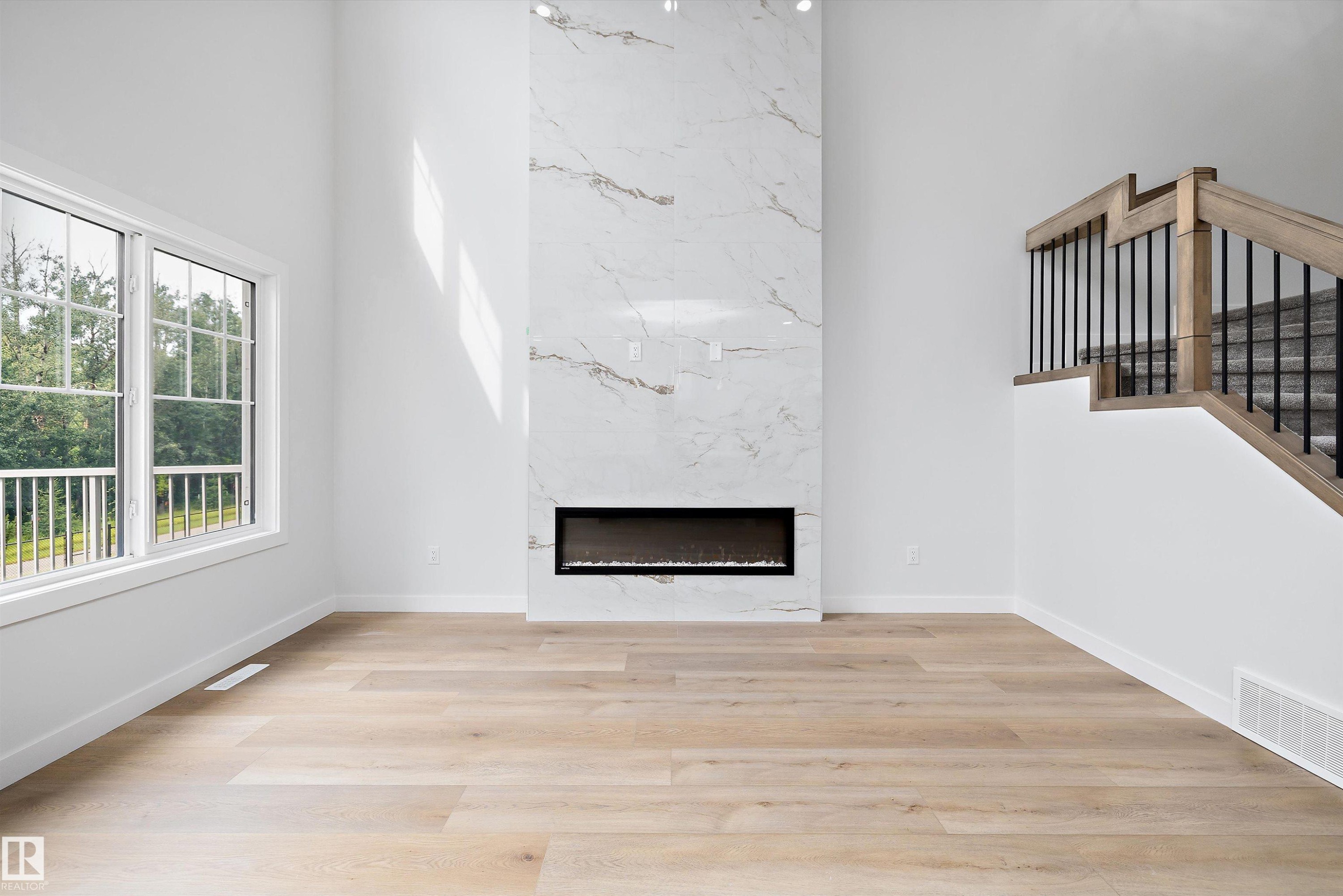 Two-story living area featuring a full-height fireplace with a linear insert and light-toned surround, wood-finish flooring, a contemporary staircase with iron balusters and wood handrails, and a large multi-pane window - 919 Elderberry Landing Nw, Edmonton, AB - Indoor With Fireplace