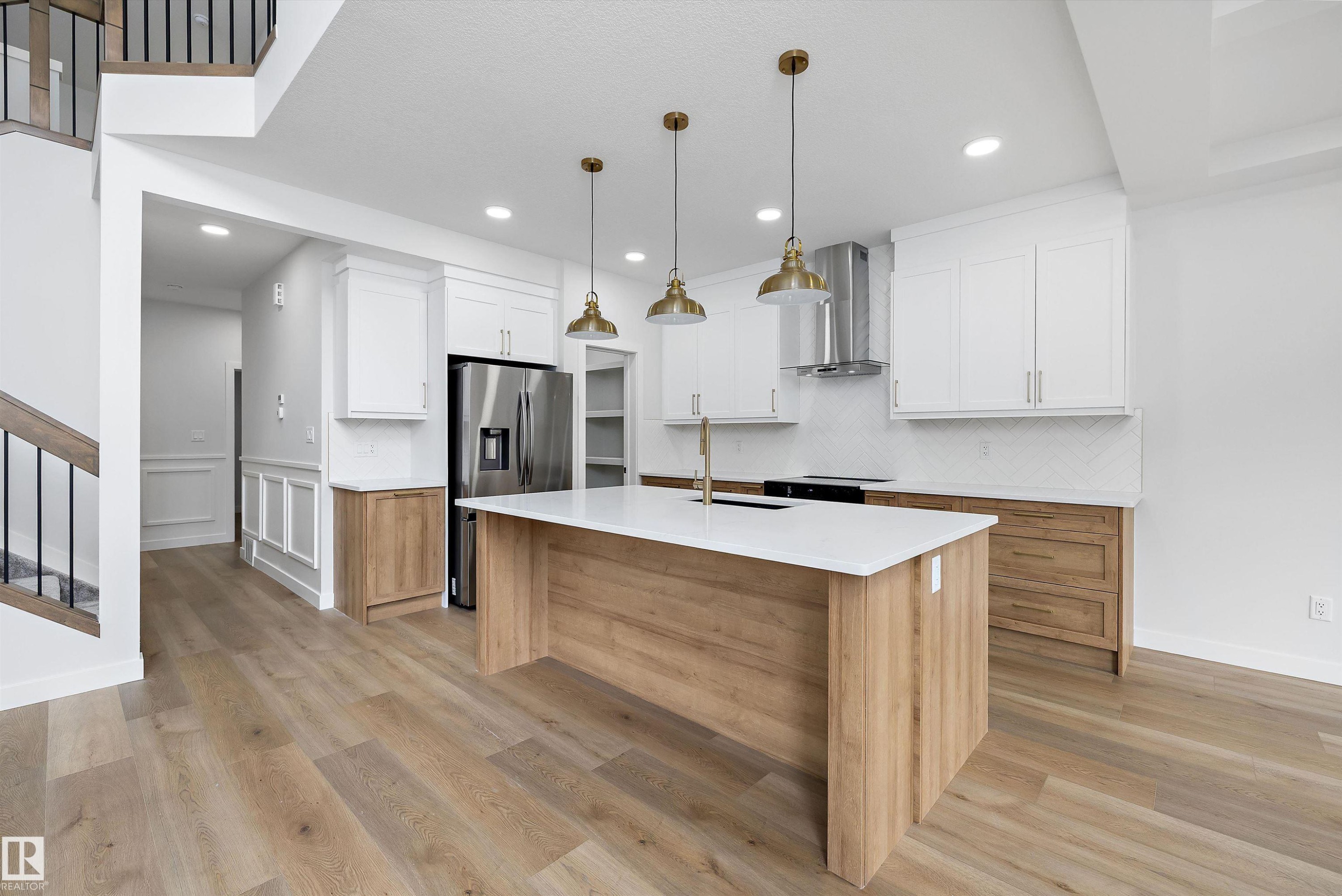 Open-concept kitchen featuring an expansive center island with a white countertop, brass pendant lighting, wood-finish flooring, white upper cabinetry, and stainless steel appliances - 919 Elderberry Landing Nw, Edmonton, AB - Indoor Photo Showing Kitchen With Upgraded Kitchen