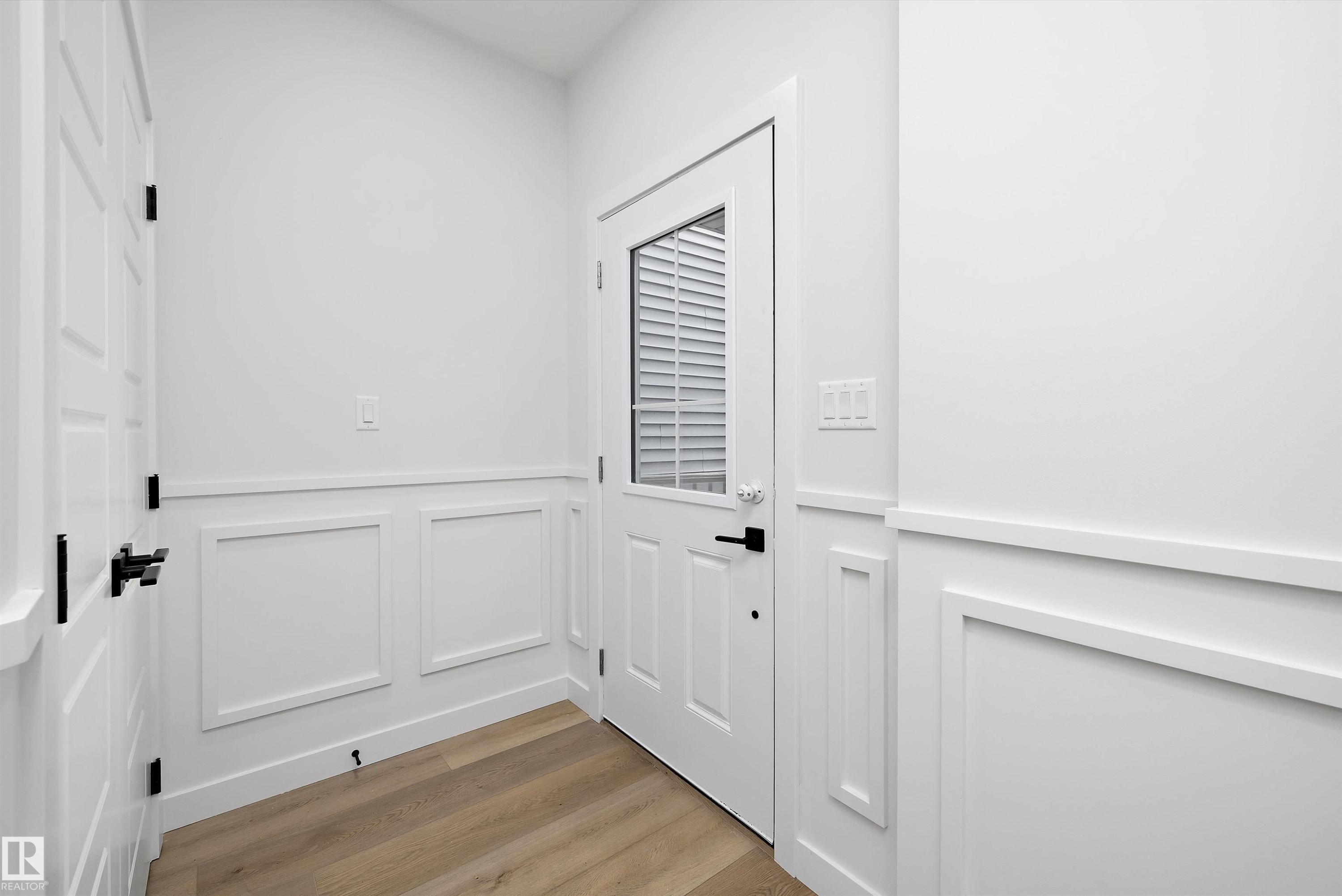 Entryway featuring wainscoting with decorative paneling, wood-finish flooring, a white paneled door with integrated blinds, and matte black hardware - 919 Elderberry Landing Nw, Edmonton, AB - Indoor Photo Showing Other Room