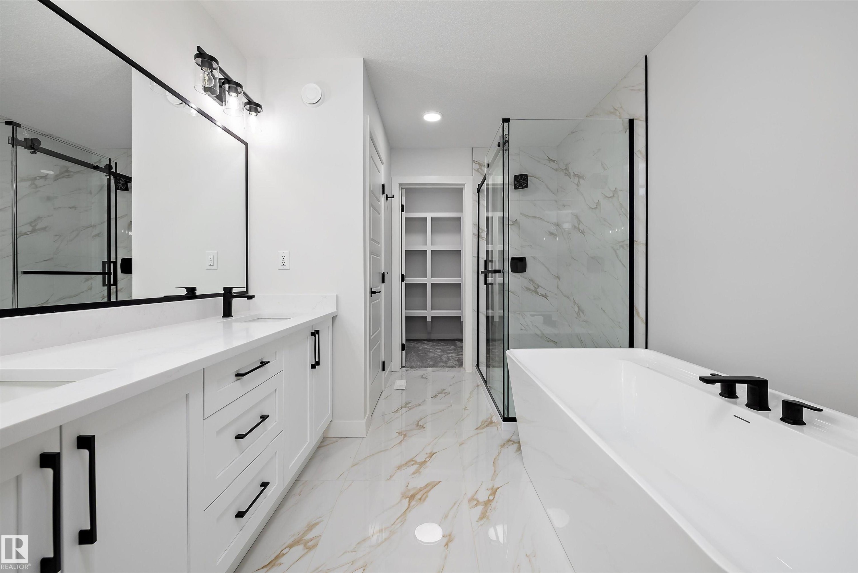 Modern bathroom featuring a dual vanity with white cabinetry and countertops, a freestanding soaking tub with matte black fixtures, a walk-in shower with clear glass enclosure and marble-patterned tile, and polished floor tiles with gold veining - 919 Elderberry Landing Nw, Edmonton, AB - Indoor Photo Showing Bathroom