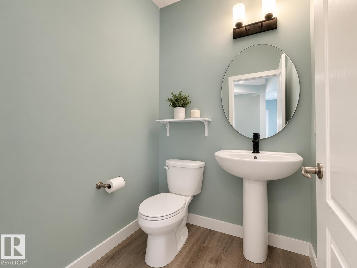 Bathroom featuring a white pedestal sink with a matte black faucet, a circular mirror, and a two-bulb vanity light - #56 804 Welsh Dr Sw, Edmonton, AB - Indoor Photo Showing Bathroom