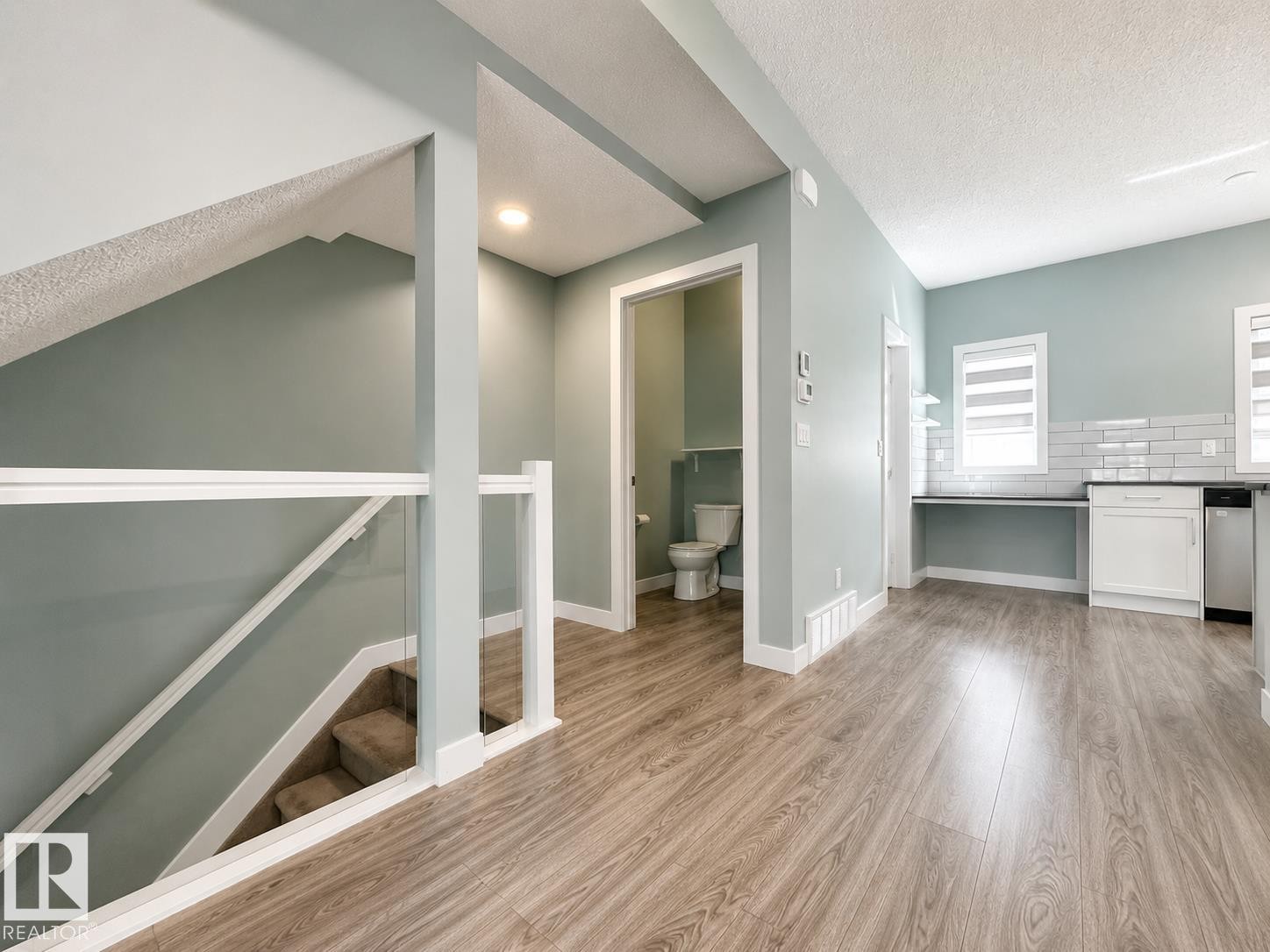 Wood-finish flooring throughout, featuring a glass-paneled staircase with white railings, recessed lighting, a built-in desk area, white cabinetry, and subway tile backsplash - #56 804 Welsh Dr Sw, Edmonton, AB - Indoor