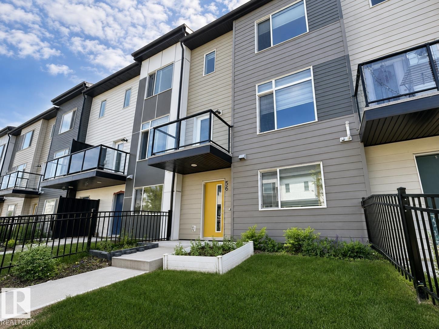 Modern townhome exterior with mixed material siding, multiple balconies featuring glass railings, a vibrant yellow front door, and a manicured lawn - #56 804 Welsh Dr Sw, Edmonton, AB - Outdoor With Balcony