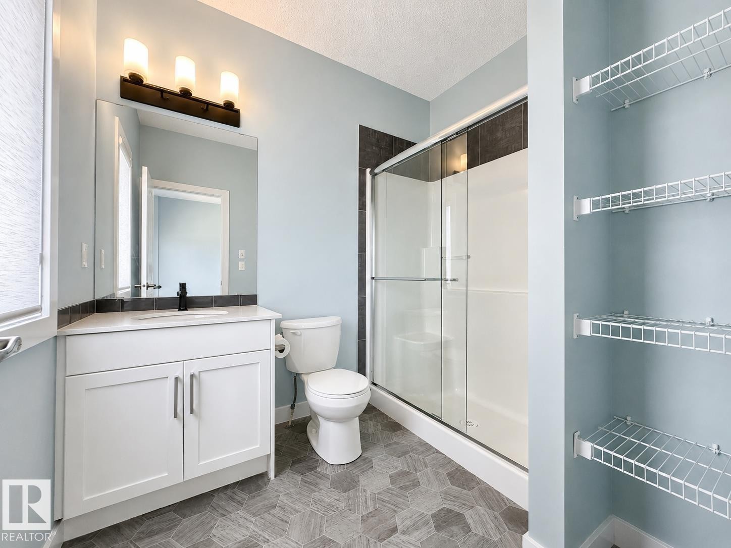 Bathroom featuring a white vanity with an undermount sink, a shower with sliding glass doors, hexagonal floor tiling, and wire shelving - #56 804 Welsh Dr Sw, Edmonton, AB - Indoor Photo Showing Bathroom