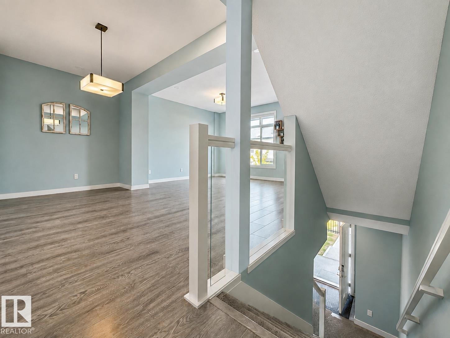 Open-concept layout with wood-finish flooring, a contemporary ceiling light fixture, and a glass-paneled stair railing - #56 804 Welsh Dr Sw, Edmonton, AB - Indoor Photo Showing Other Room
