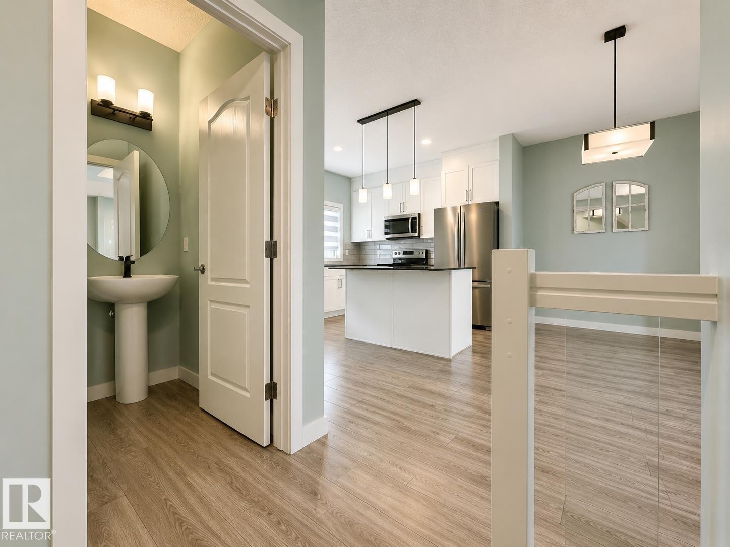 Wood-finish flooring throughout, featuring a modern pedestal sink, black wall-mounted vanity light, white Shaker-style kitchen cabinetry, and stainless steel appliances - #56 804 Welsh Dr Sw, Edmonton, AB - Indoor Photo Showing Kitchen