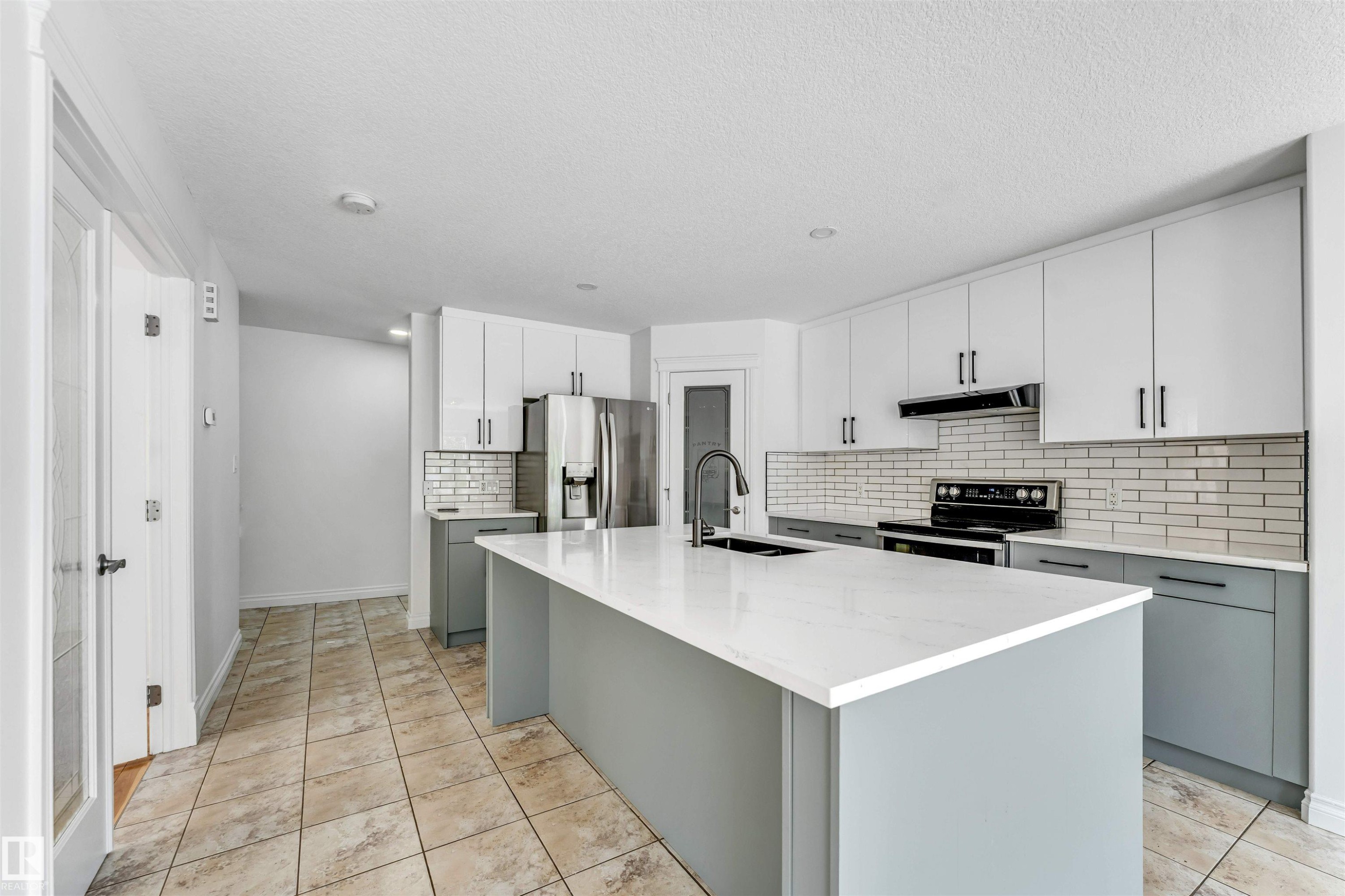 Modern kitchen featuring a large central island with a solid surface countertop, two-tone cabinetry with white upper and gray lower cabinets, full tile backsplash, stainless steel appliances, and tile flooring - 16205 55 Street Nw, Edmonton, AB - Indoor Photo Showing Kitchen With Upgraded Kitchen
