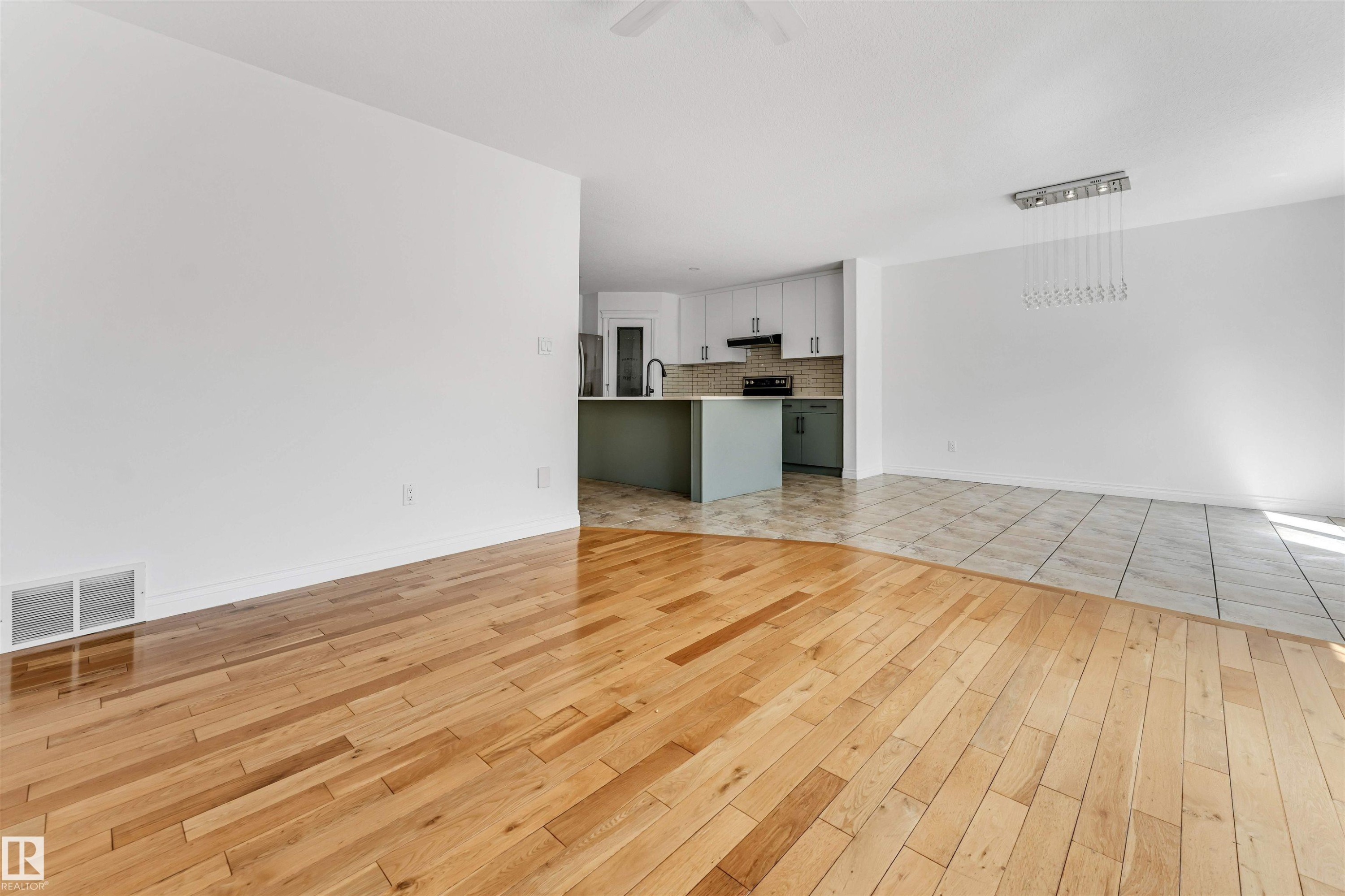 Open-concept living space featuring wood-finish flooring, a contemporary linear chandelier, and a kitchen with two-tone cabinetry, a central island, and a tile backsplash - 16205 55 Street Nw, Edmonton, AB - Indoor