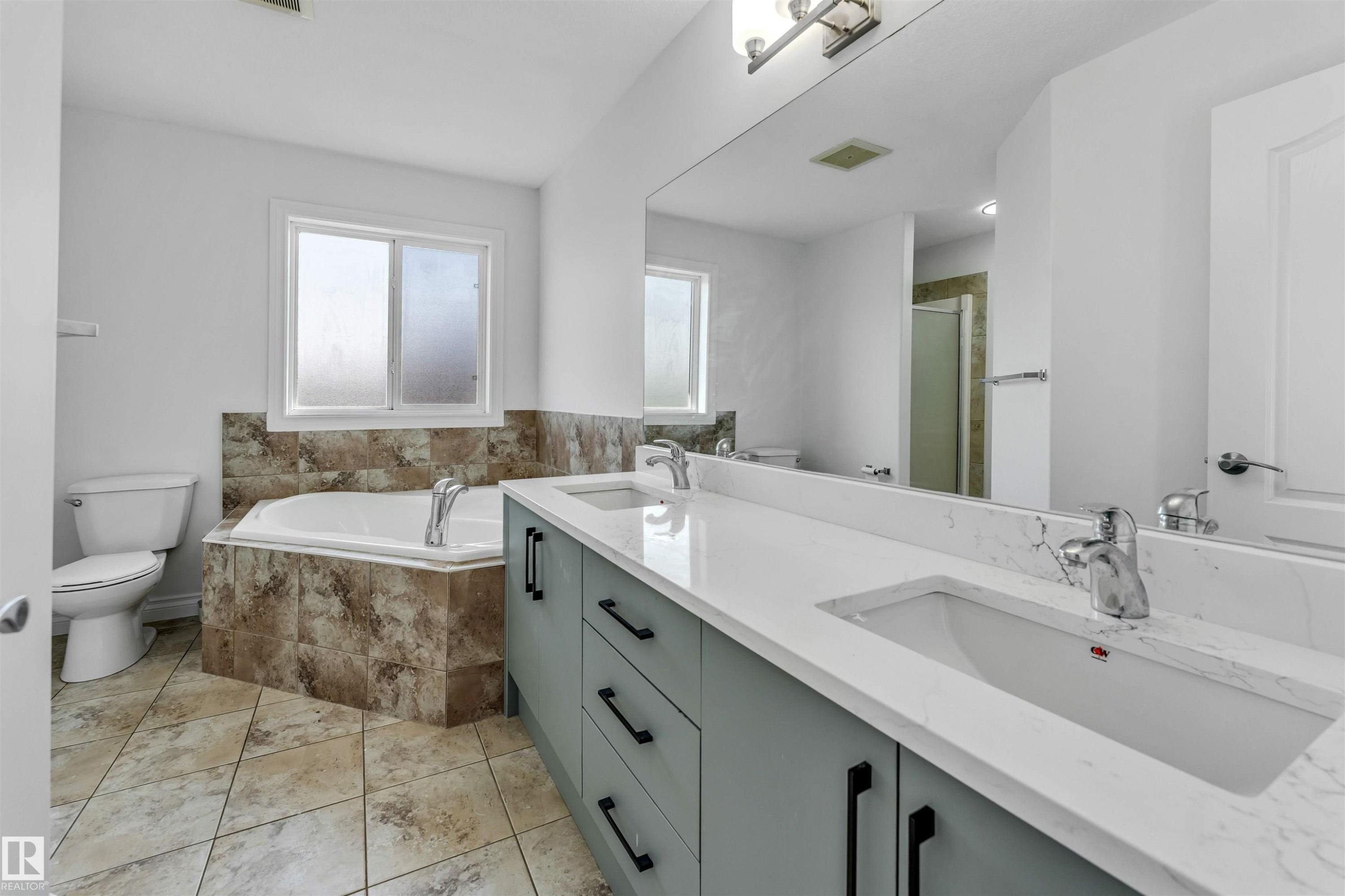 Bathroom featuring a double vanity with light gray cabinetry and white countertops, a large mirror, a built-in corner bathtub with tiled surround, and patterned floor tiling - 16205 55 Street Nw, Edmonton, AB - Indoor Photo Showing Bathroom