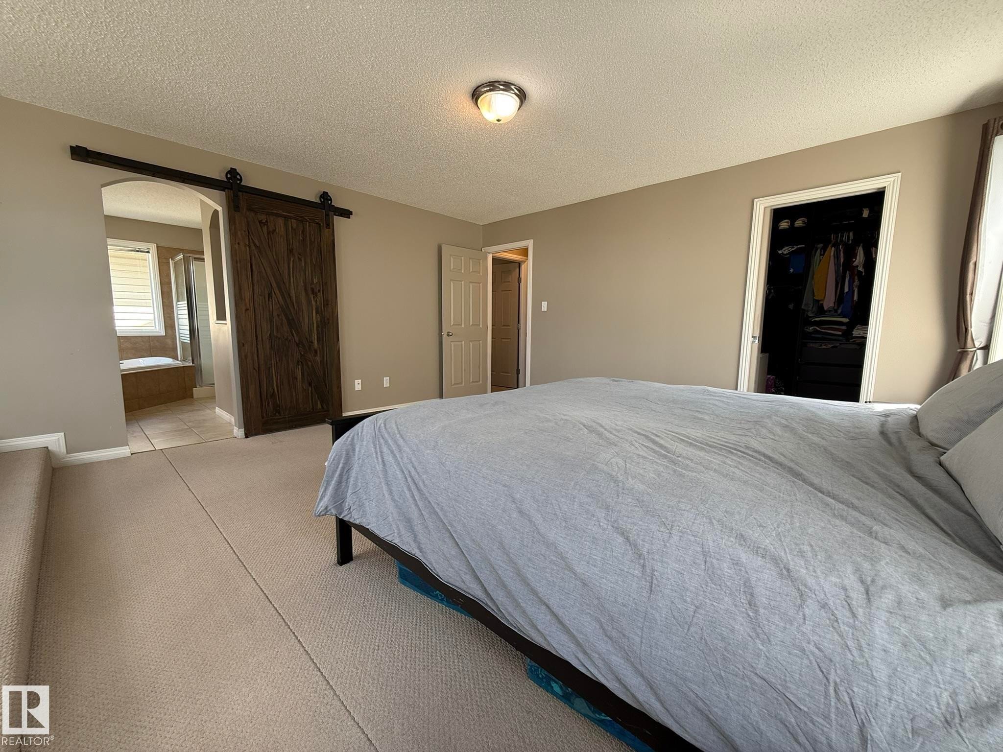 Spacious carpeted room featuring a rustic wood barn door, recessed closet, and an arched entry to a tiled area with a built-in tub - 1820 Robertson Crescent Sw, Edmonton, AB - Indoor Photo Showing Bedroom