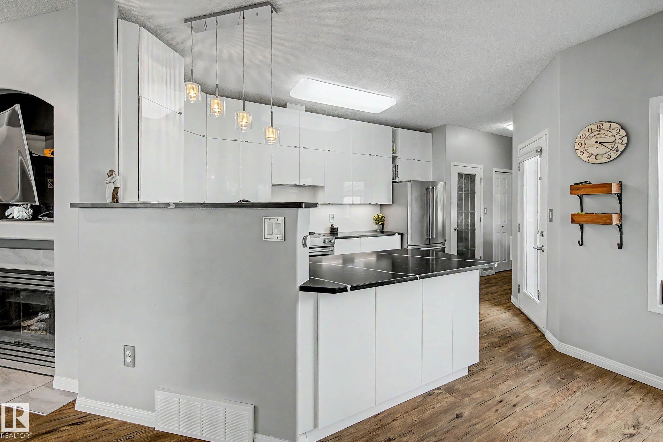 Modern kitchen featuring glossy white cabinetry, dark stone countertops, stainless steel appliances, a breakfast bar, and wood-finish flooring - 9130 100A Avenue Nw, Edmonton, AB - Indoor Photo Showing Kitchen