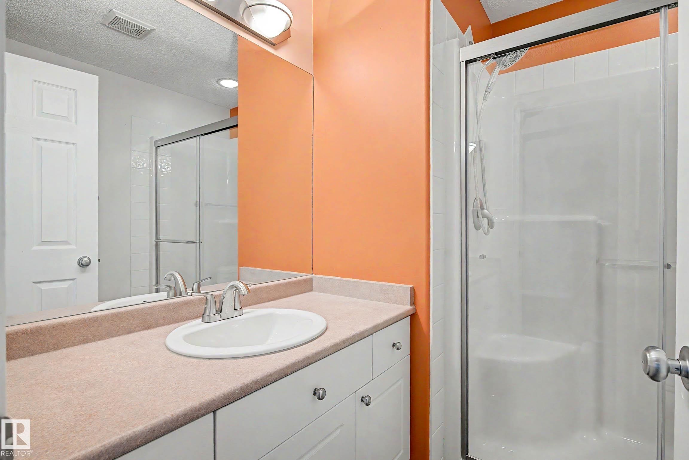 Bathroom featuring a vanity with an integrated sink and light-toned countertop, white cabinetry with silver hardware, a large wall-mounted mirror, and a shower with a sliding glass enclosure - 9130 100A Avenue Nw, Edmonton, AB - Indoor Photo Showing Bathroom