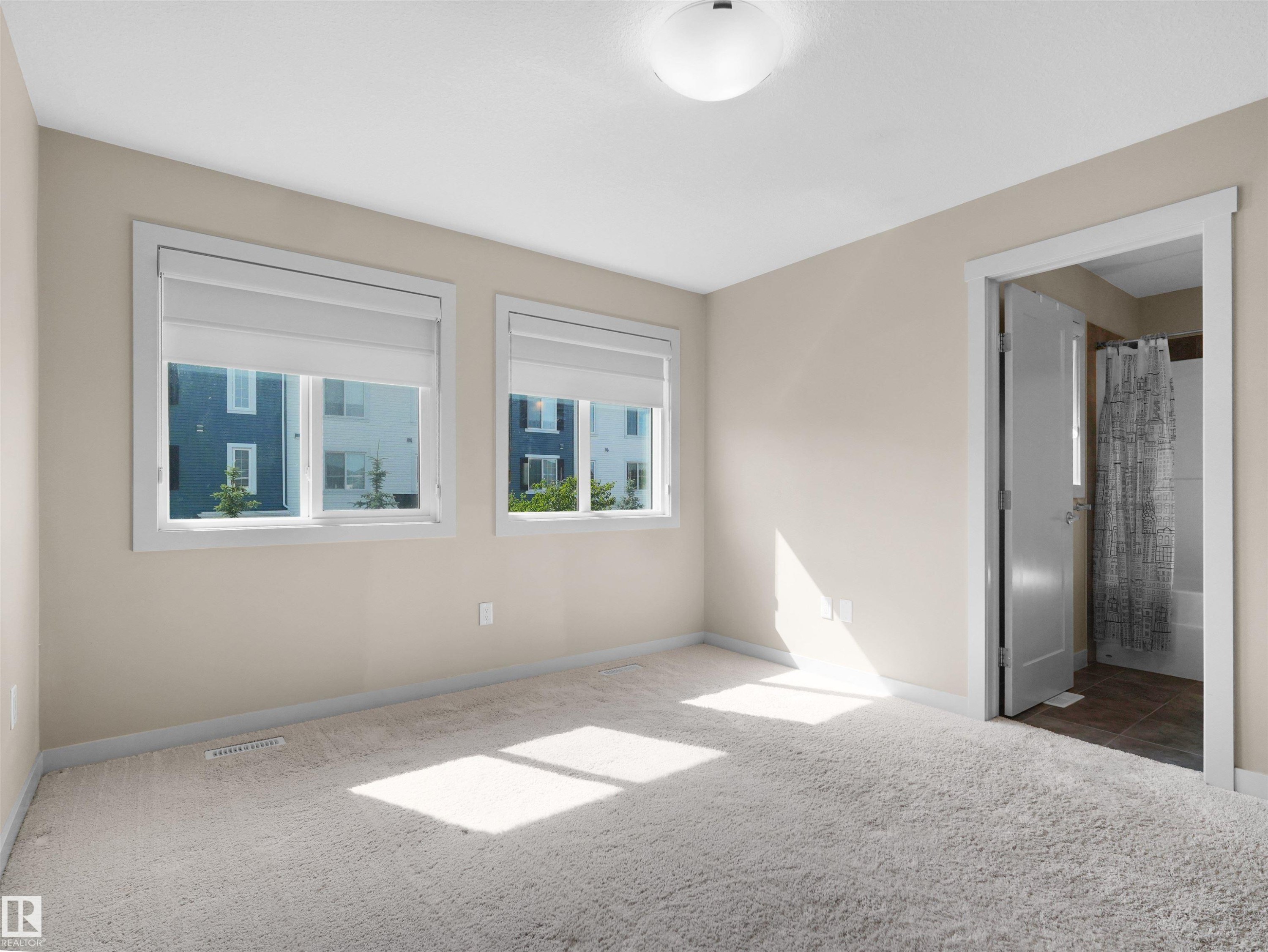 Carpeted room featuring two windows with roller blinds, a ceiling-mounted light fixture, and a doorway revealing a partial bathroom with a shower-tub combination and tiled flooring - 4487 Annett Common Sw, Edmonton, AB - Indoor