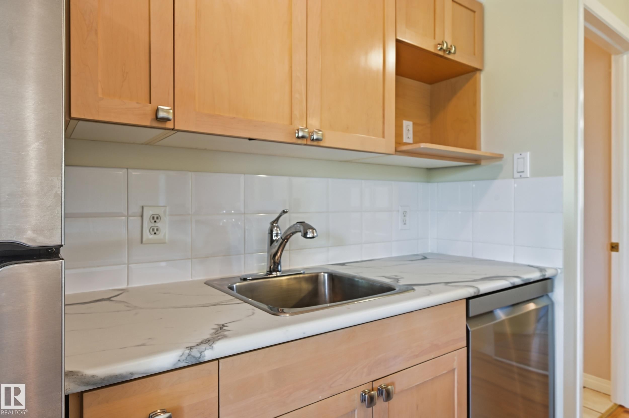 Kitchen features light wood cabinetry, white tile backsplash, stainless steel sink with chrome faucet, and marbling-patterned countertops - #433 11217 103 Av Nw, Edmonton, AB - Indoor Photo Showing Kitchen