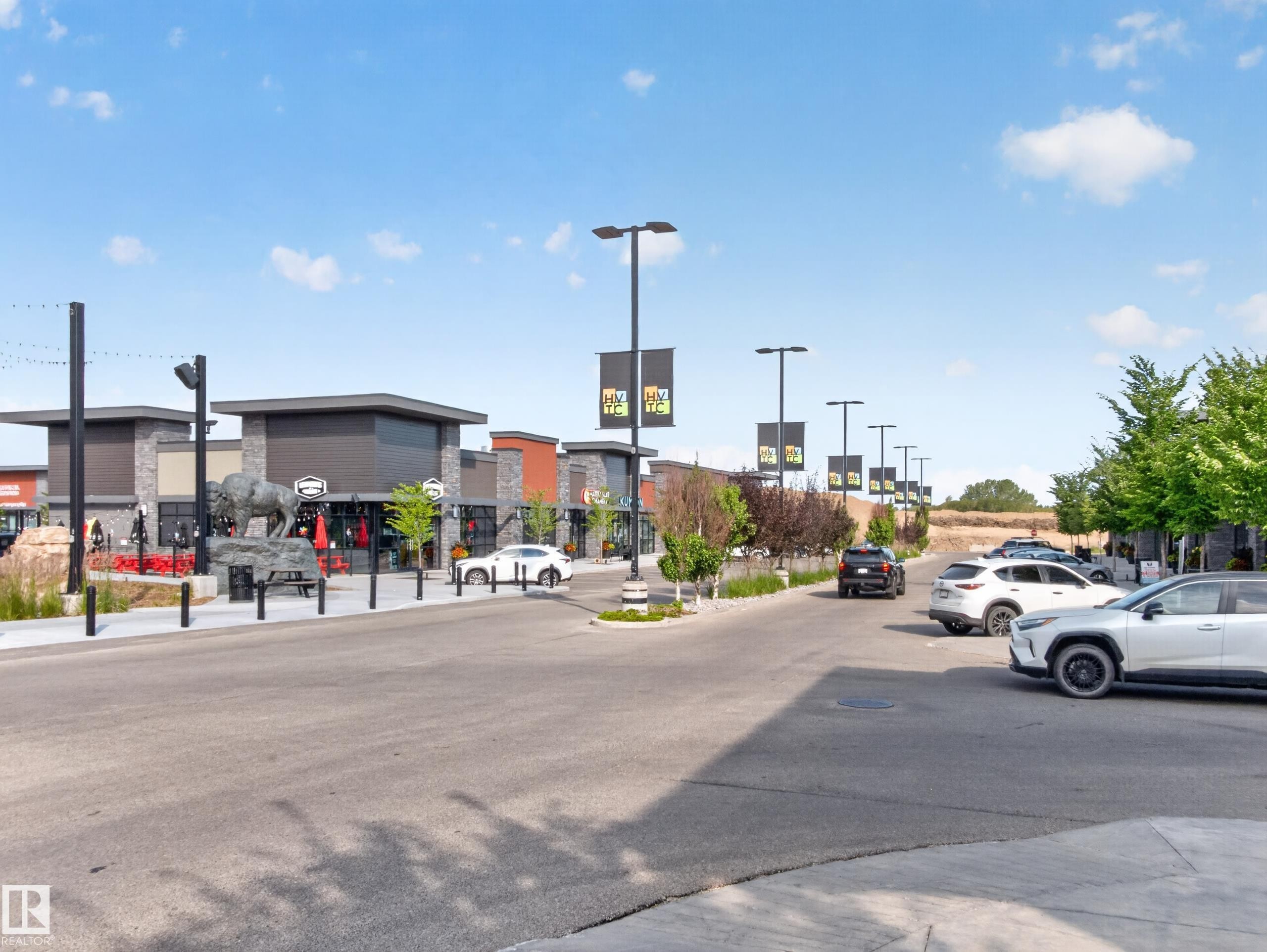 Modern commercial complex featuring contemporary architecture with stone and panel facades, multiple storefronts, a prominent public art sculpture, street lighting, and manicured landscaping - #217 12045 22 Av Sw, Edmonton, AB - Outdoor