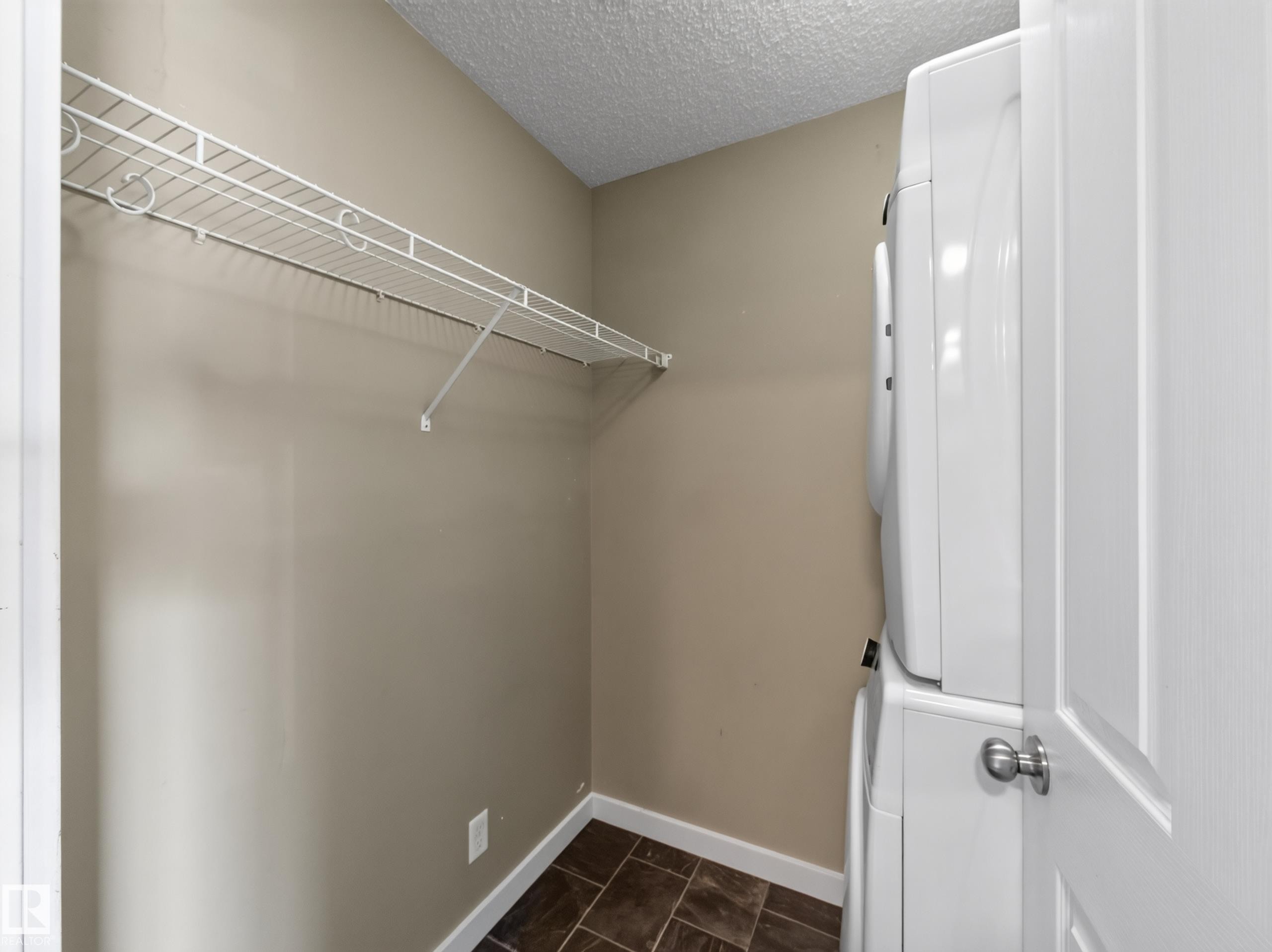 Dedicated laundry area featuring a stacked washer and dryer, wire shelving, textured ceiling, neutral wall paint, and dark tile flooring - #217 12045 22 Av Sw, Edmonton, AB - Indoor With Storage