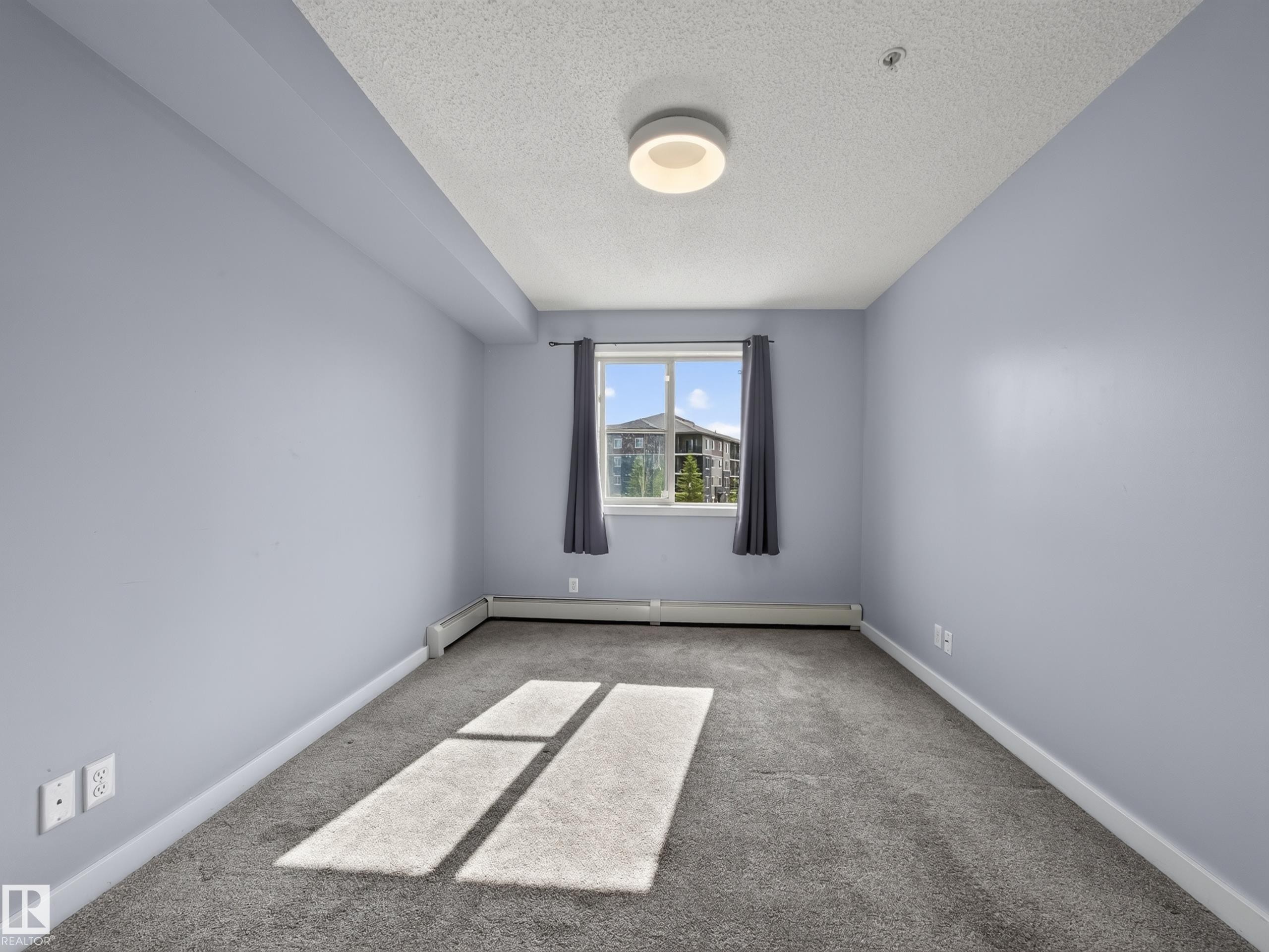Spacious room featuring light blue walls, gray carpet, and a window offering exterior views - #217 12045 22 Av Sw, Edmonton, AB - Indoor Photo Showing Other Room